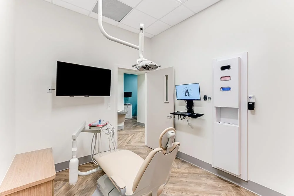 Dentist's examination room with dental chair, wall-mounted monitor, medical supplies, and a door leading to an adjacent room.