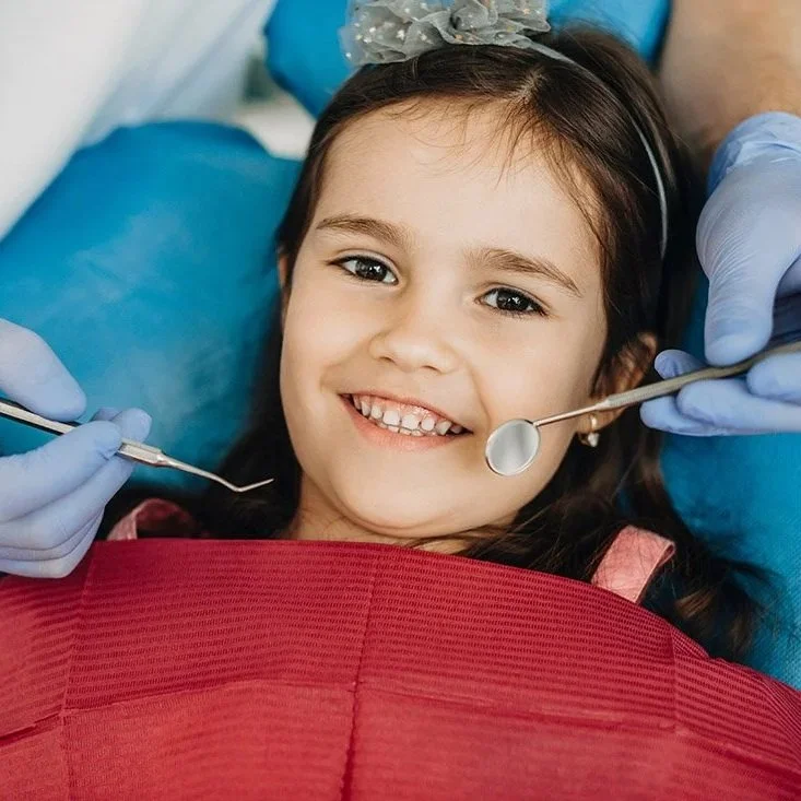 Young girl smiling in dental chair during checkup, with dentist's gloved hands holding dental tools near her face
