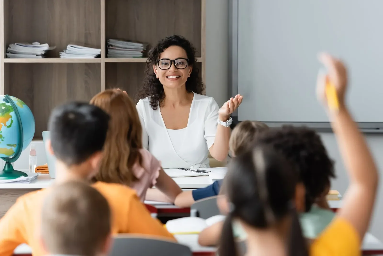 A teacher sitting at a desk in front of a classroom with students, some of whom have raised their hands, in a classroom with shelves and a globe.