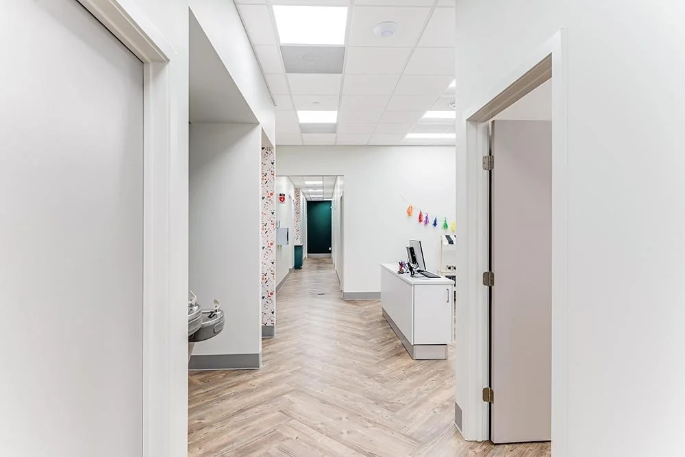 Empty hallway in a modern dentist office with white walls, wood-patterned flooring, a reception desk with a computer, and colorful decorations on the wall.