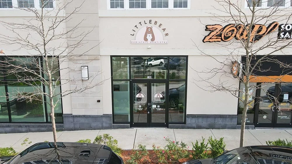 Street view showing two leafless trees in front of a commercial building housing Littlebeard Pediatric Dentistry and Zoup. There are parked cars along the sidewalk and large glass windows.