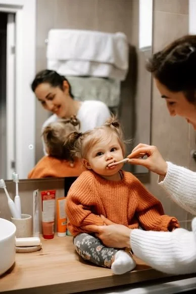A toddler girl sitting on a bathroom counter while an adult woman brushed her teeth.