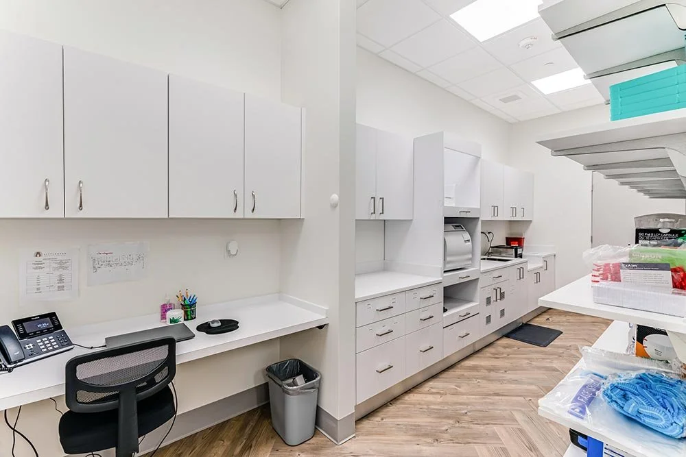 A clean, organized dental supply room with white cabinets, a counter, a telephone, a chair, and shelves holding medical supplies and equipment.