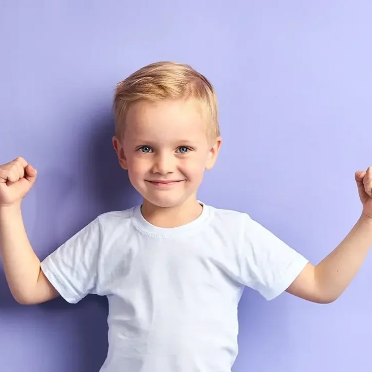 A young boy with blonde hair and blue eyes smiling and flexing his arms in a white t-shirt against a light purple background.