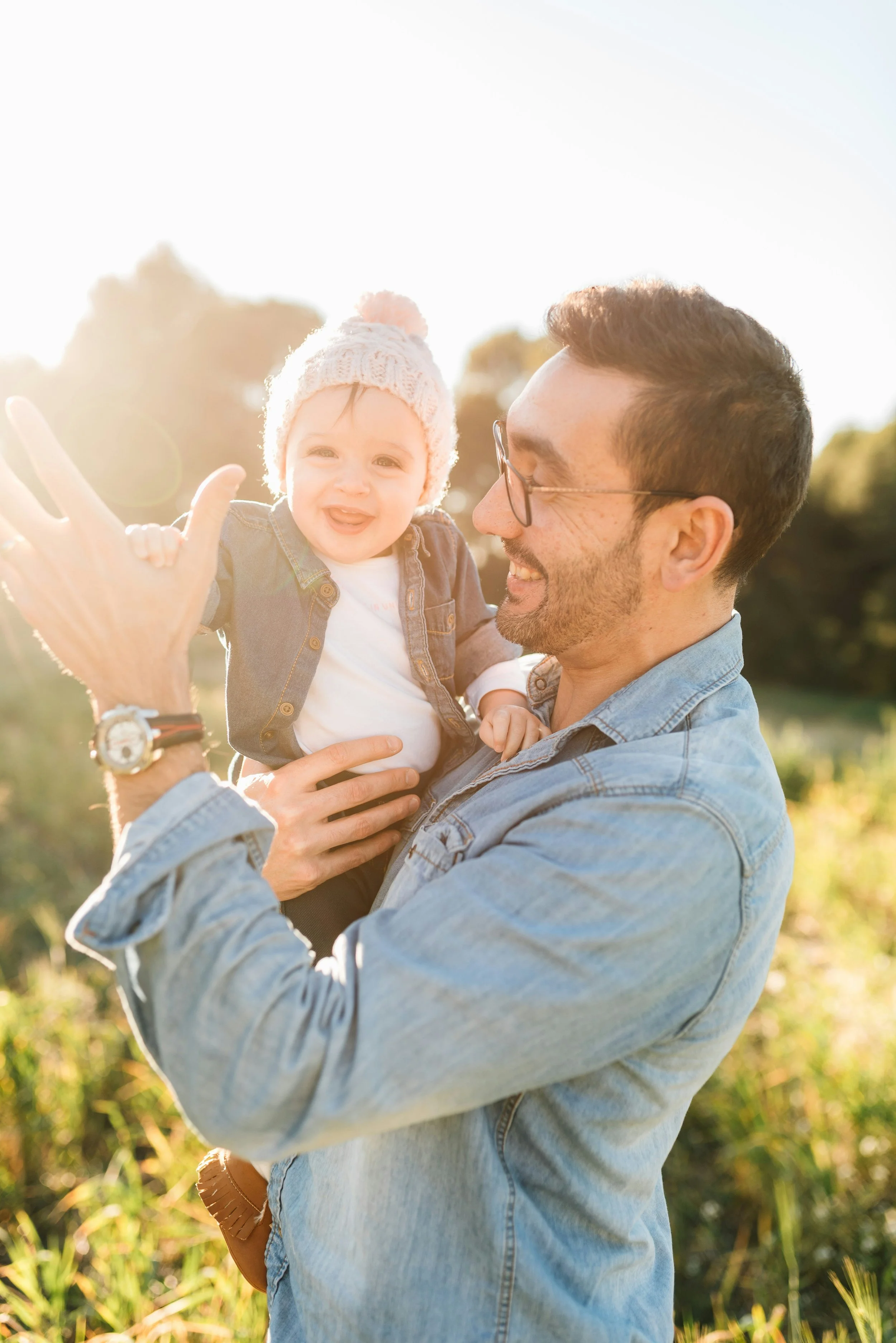 A man with glasses and a beard holding a smiling baby girl outdoors during sunset. The baby wears a pink knit hat with a pom-pom and a denim jacket, while the man wears a light blue denim shirt. They are surrounded by greenery and appear joyful.