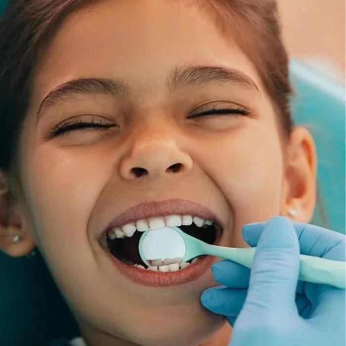 A young girl at the dentist having her teeth checked, with her eyes closed and mouth open, while a dental hygienist holds a mirror to examine her teeth.