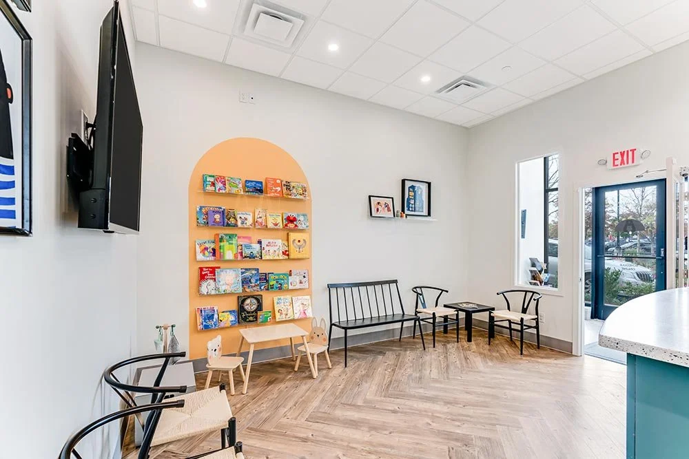 Interior of a waiting room with a bookshelf filled with children's books, two chairs with black frames, a black bench, and a small table, a wall-mounted TV, and an exit door with a window.