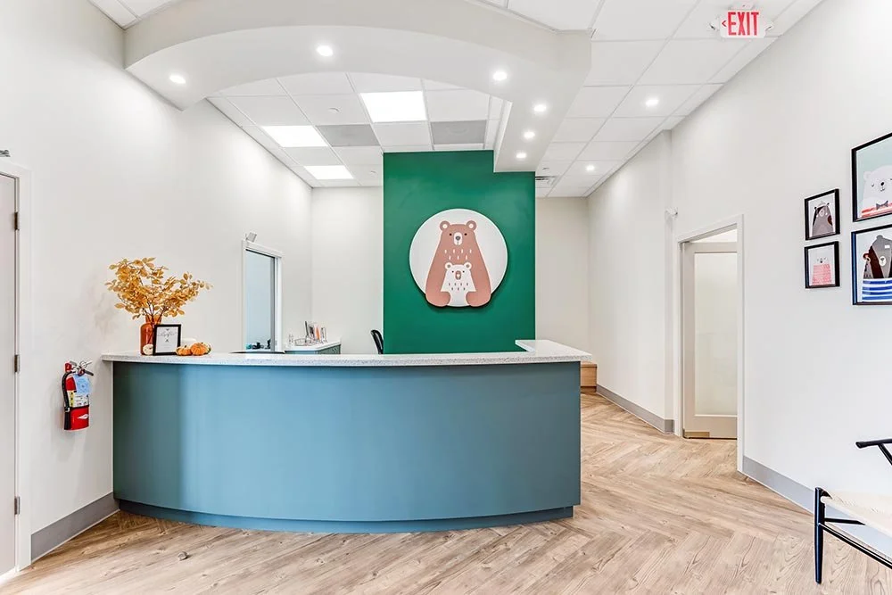 Reception area of a dentist office with a blue curved counter, a green wall with a bear logo, framed animal artwork, a fire extinguisher, and a decorative plant.