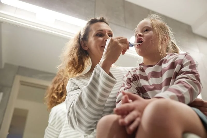 A woman with long curly hair helping a young girl brush her teeth in a bathroom.