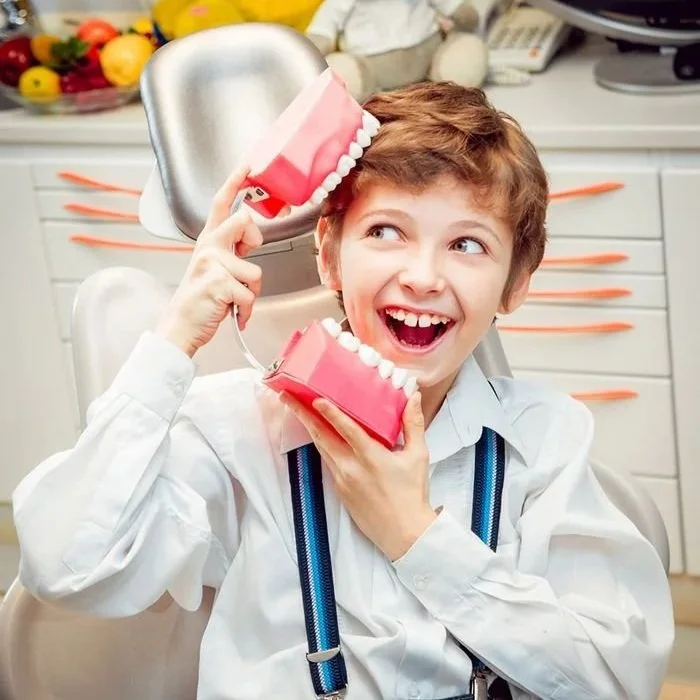 A boy with brown hair smiling and holding a large plastic model of dentures and a toothbrush.