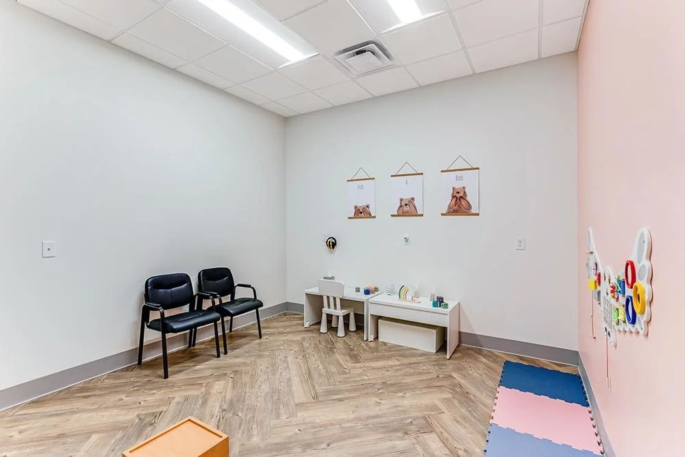Empty waiting room with three black chairs, a small table, and colorful toys on the pink wall. There are three framed pictures of dogs on the white wall.