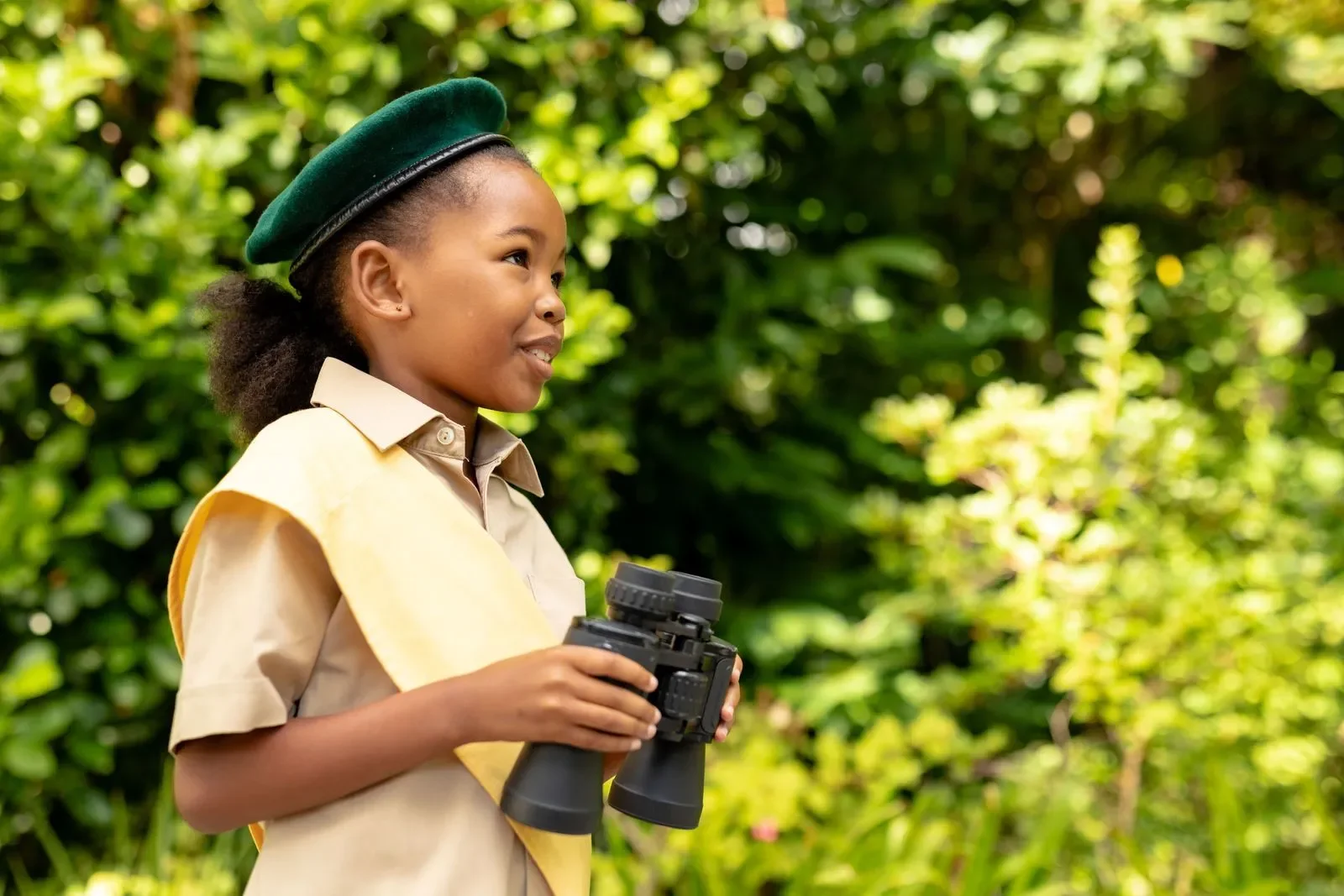 A young girl dressed in a tan uniform with a yellow sash, wearing a dark green cap, holding a pair of binoculars, standing outdoors in a lush green setting.