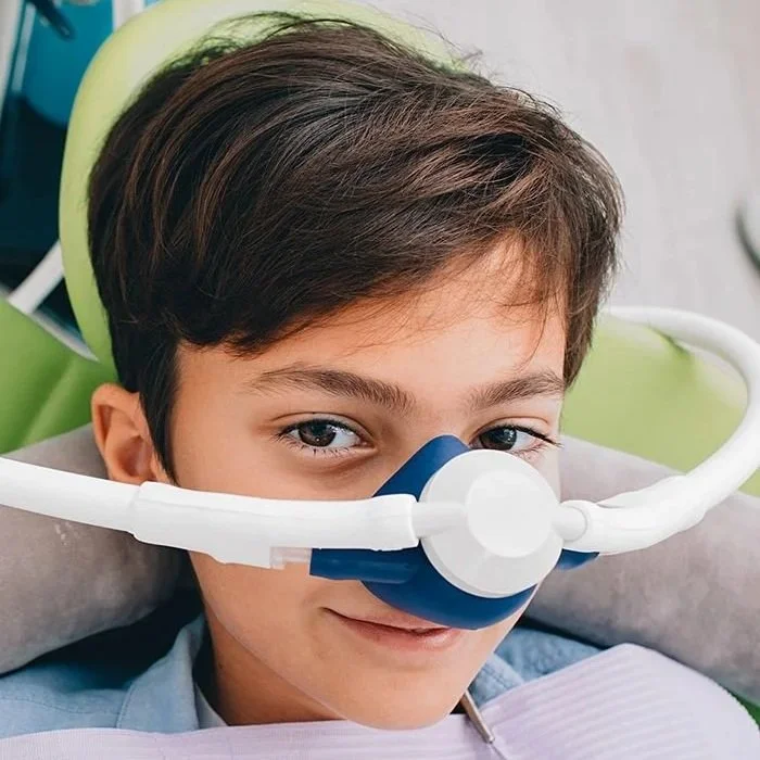 Young boy with brown hair lying in a medical bed, wearing a medical oxygen mask with a clear plastic tube. He looks at the camera with a slight smile.
