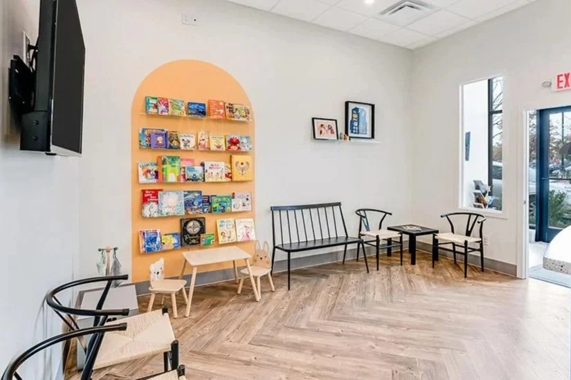 A cozy waiting room with a bookshelf filled with colorful children's books, benches, chairs, and small tables, with natural light coming in through large windows.