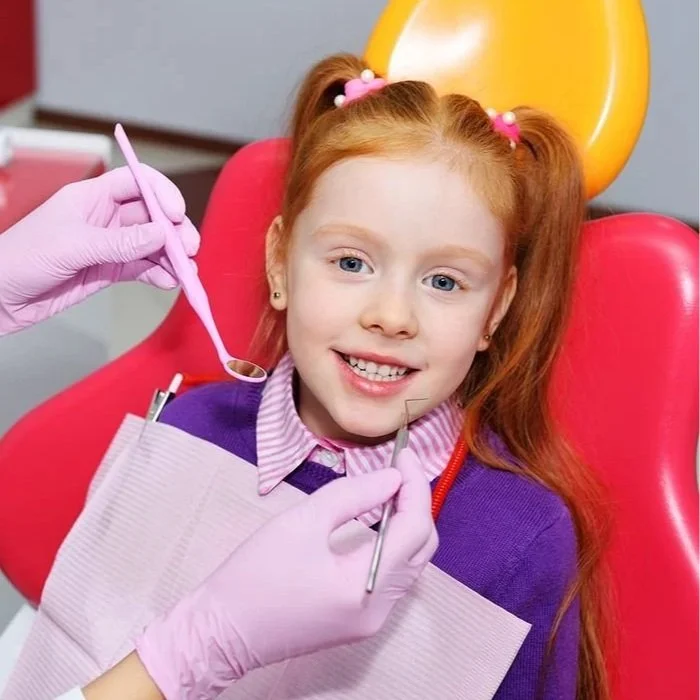 A young girl with red hair and a purple sweater sitting in a dental chair while a dentist with pink gloves examines her teeth with dental tools.