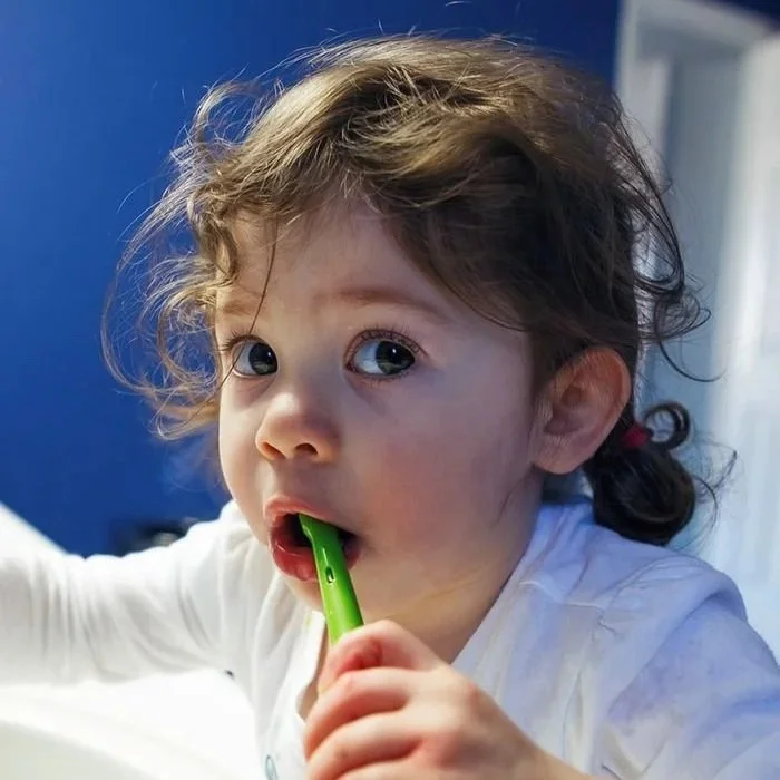 A young girl with curly hair and blue eyes biting on a green toothbrush.