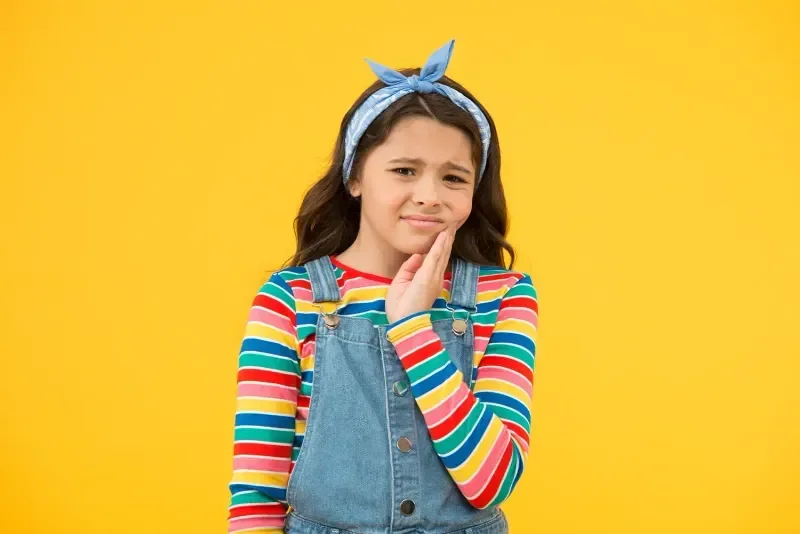 A young girl with long brown hair wearing a blue headband, a colorful striped long sleeve shirt, and denim overalls, holding her mouth with a pained expression against a yellow background.