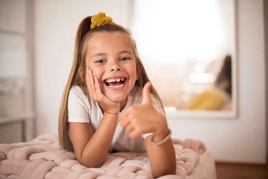 A young girl with a yellow hair bow smiling and giving a thumbs-up.