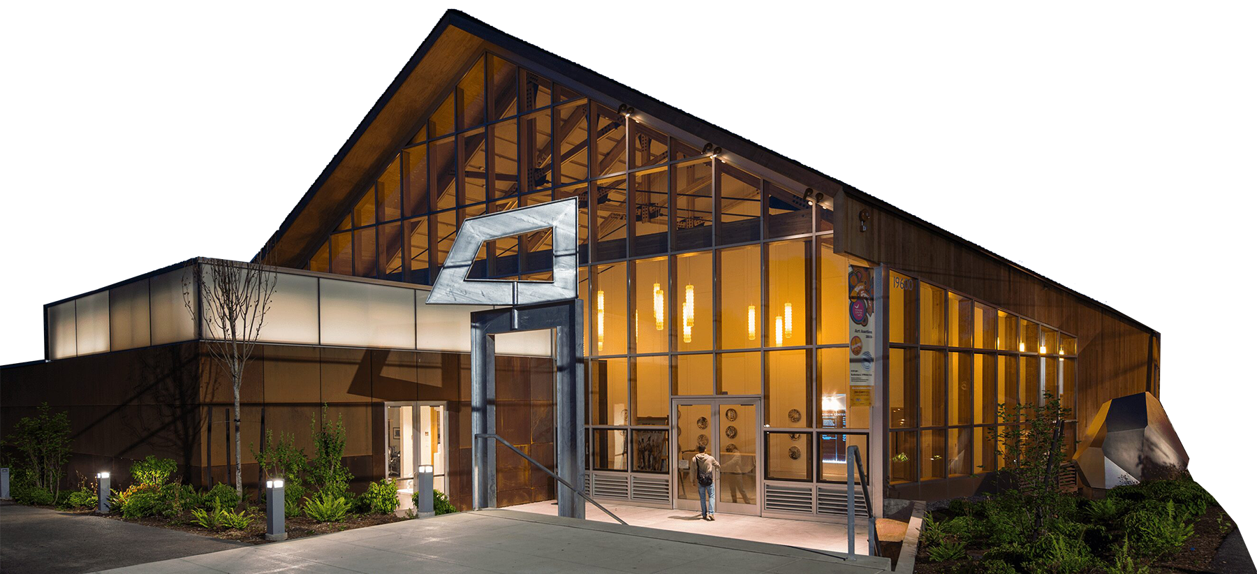 Modern building with large glass windows and wooden accents illuminated at night. A metal sculpture stands near the entrance, with landscaping and pathway lights visible.