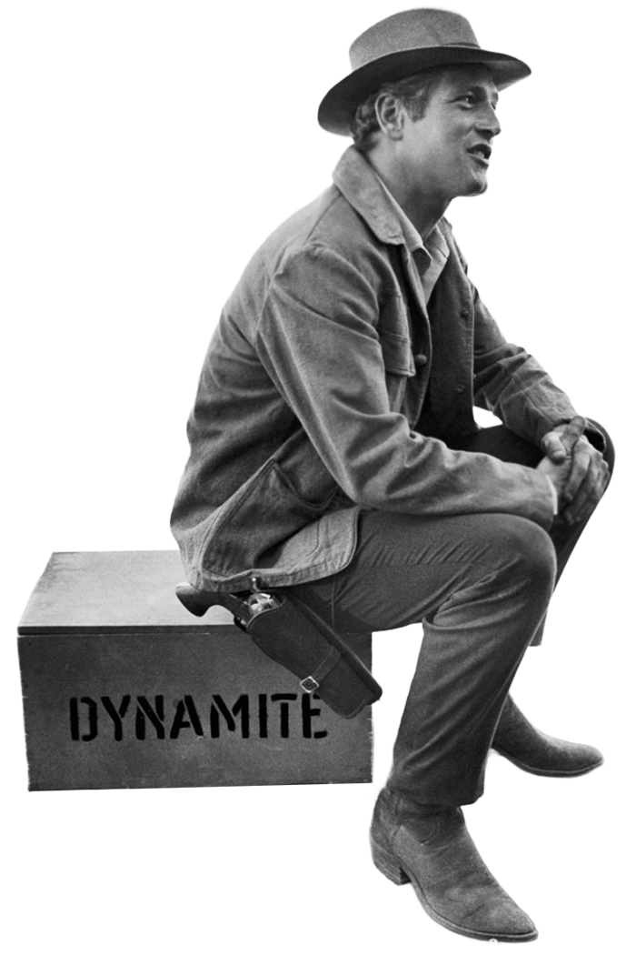 A man sitting on a wooden box labeled 'DYNAMITE' wearing a hat, denim jacket, and boots, with a handgun holstered at his side.