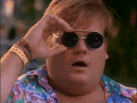 A person with red hair and sunglasses, wearing a colorful shirt, holding sunglasses up to their face, with a surprised expression.