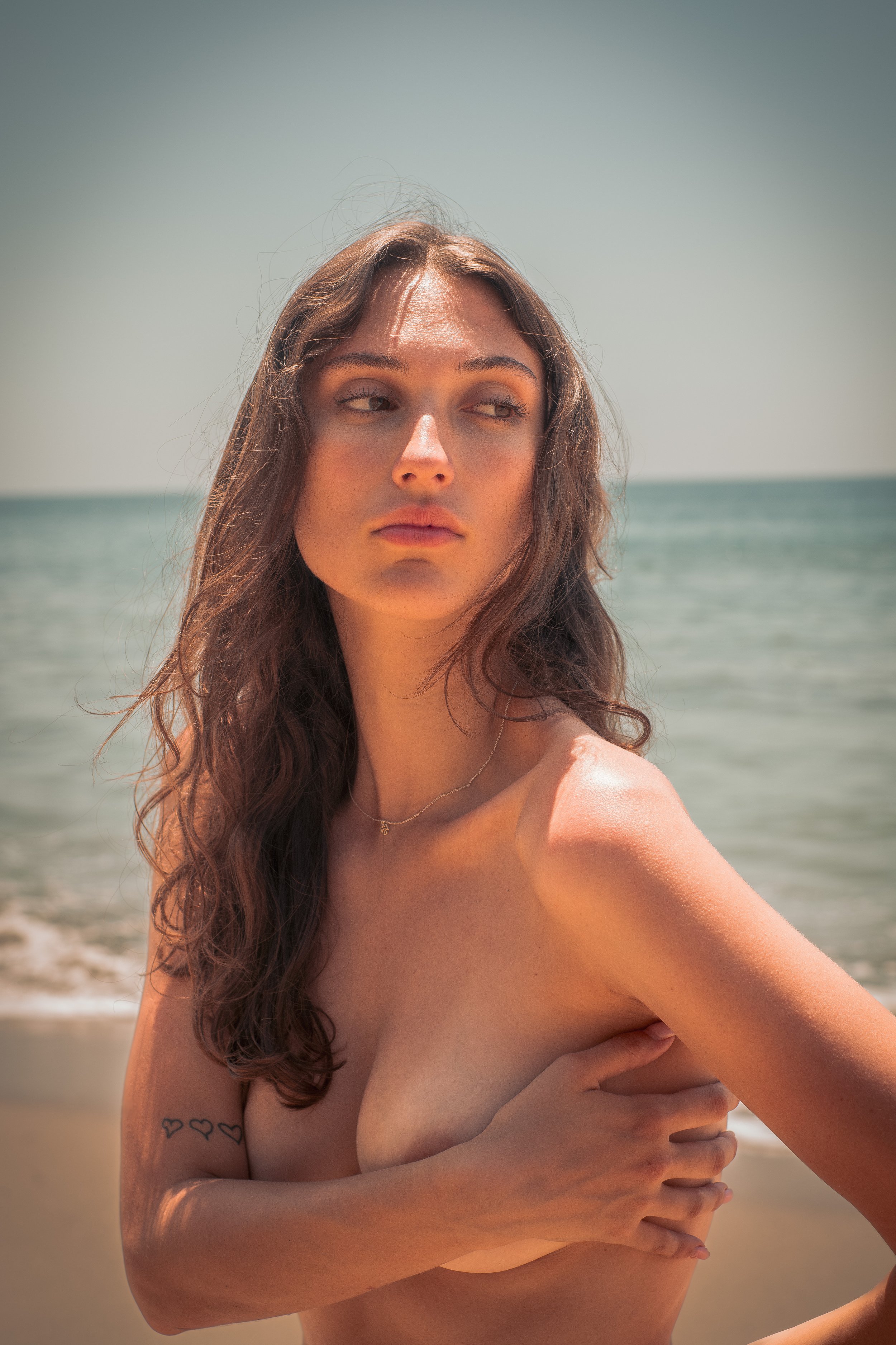 A woman with long wavy hair standing topless on the beach, holding her arm across her chest, with a tattoo of three hearts on her arm, and the ocean and sky in the background.