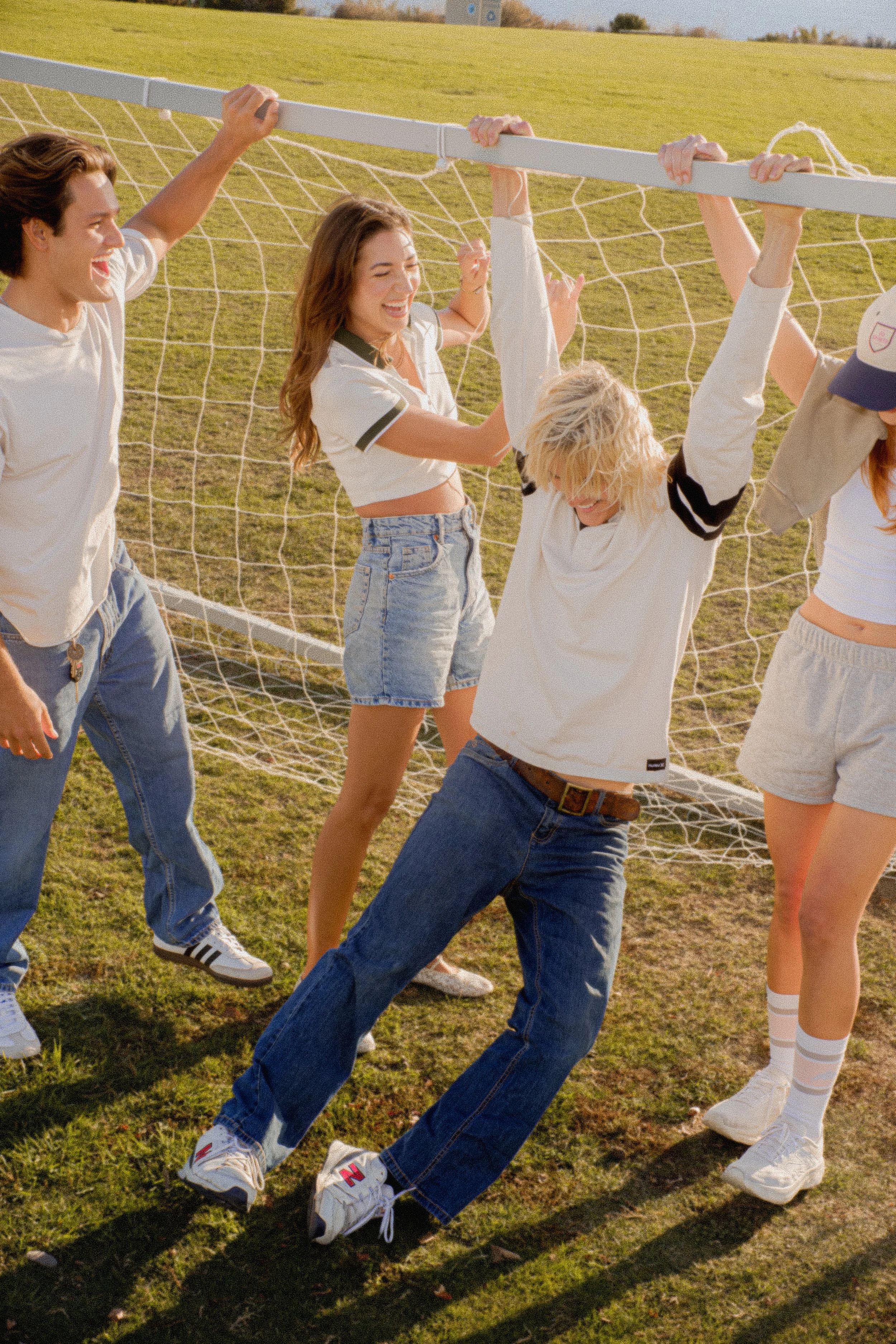 Group of young friends playing and laughing on a grassy field on a sunny day, with some holding or climbing on a soccer goalpost.