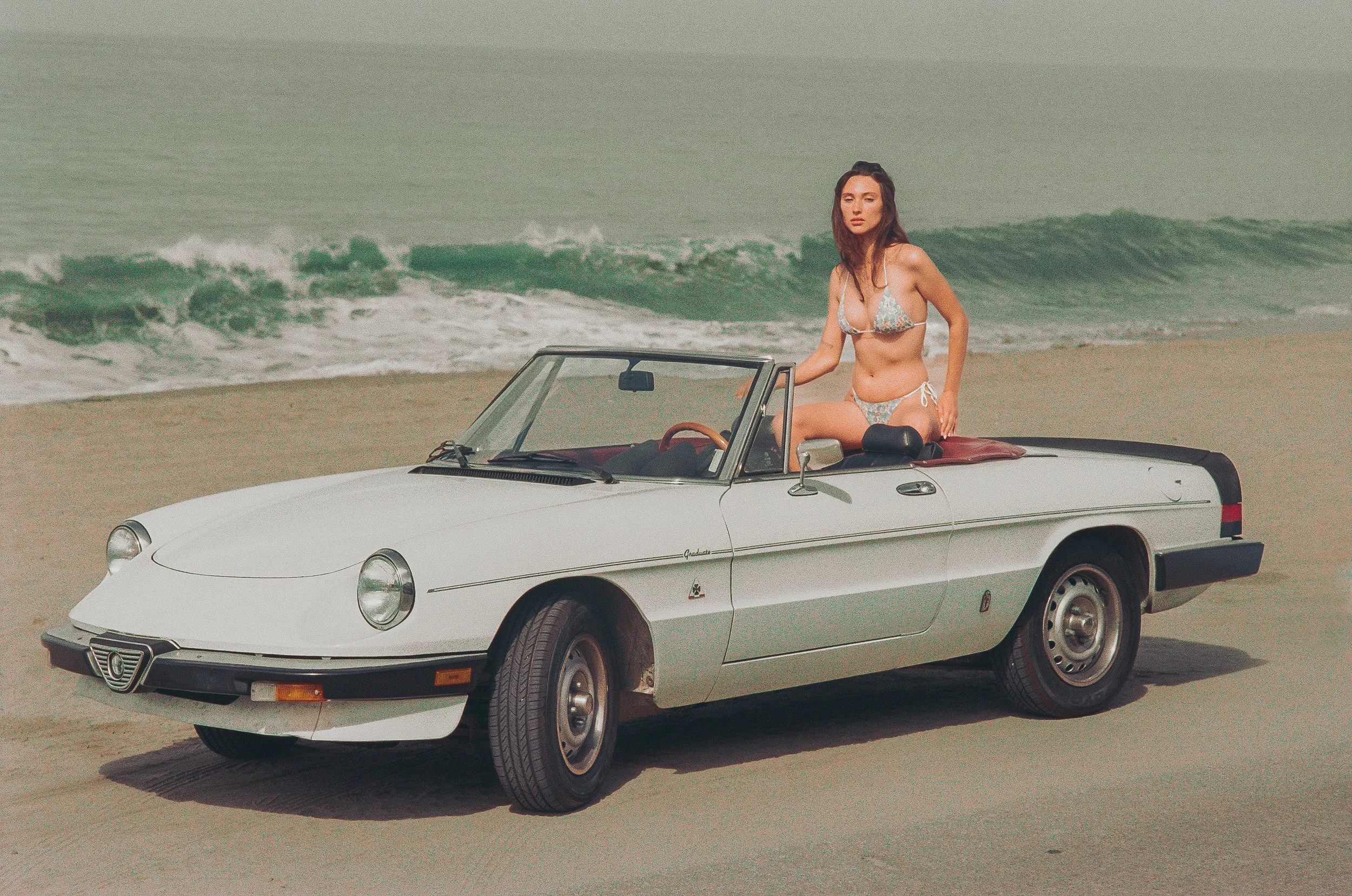 A woman in a floral bikini is sitting on the door of a vintage white convertible car on a sandy beach, with ocean waves in the background.