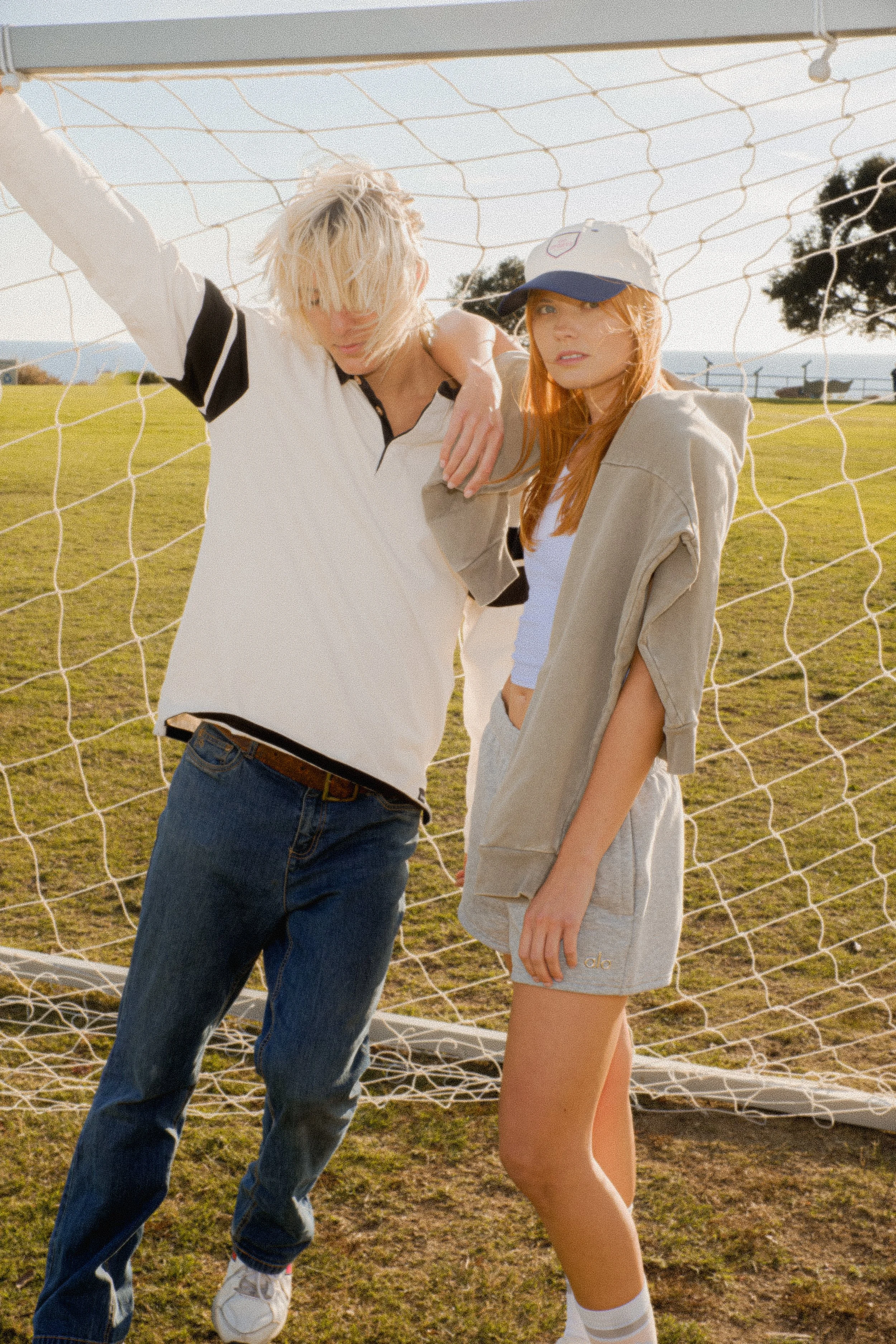 A young woman with red hair wearing a beige hoodie, shorts, and white socks, standing in front of a soccer goal with a young man with blonde hair wearing a white and black sports jersey and jeans, on a grassy field with trees in the background.