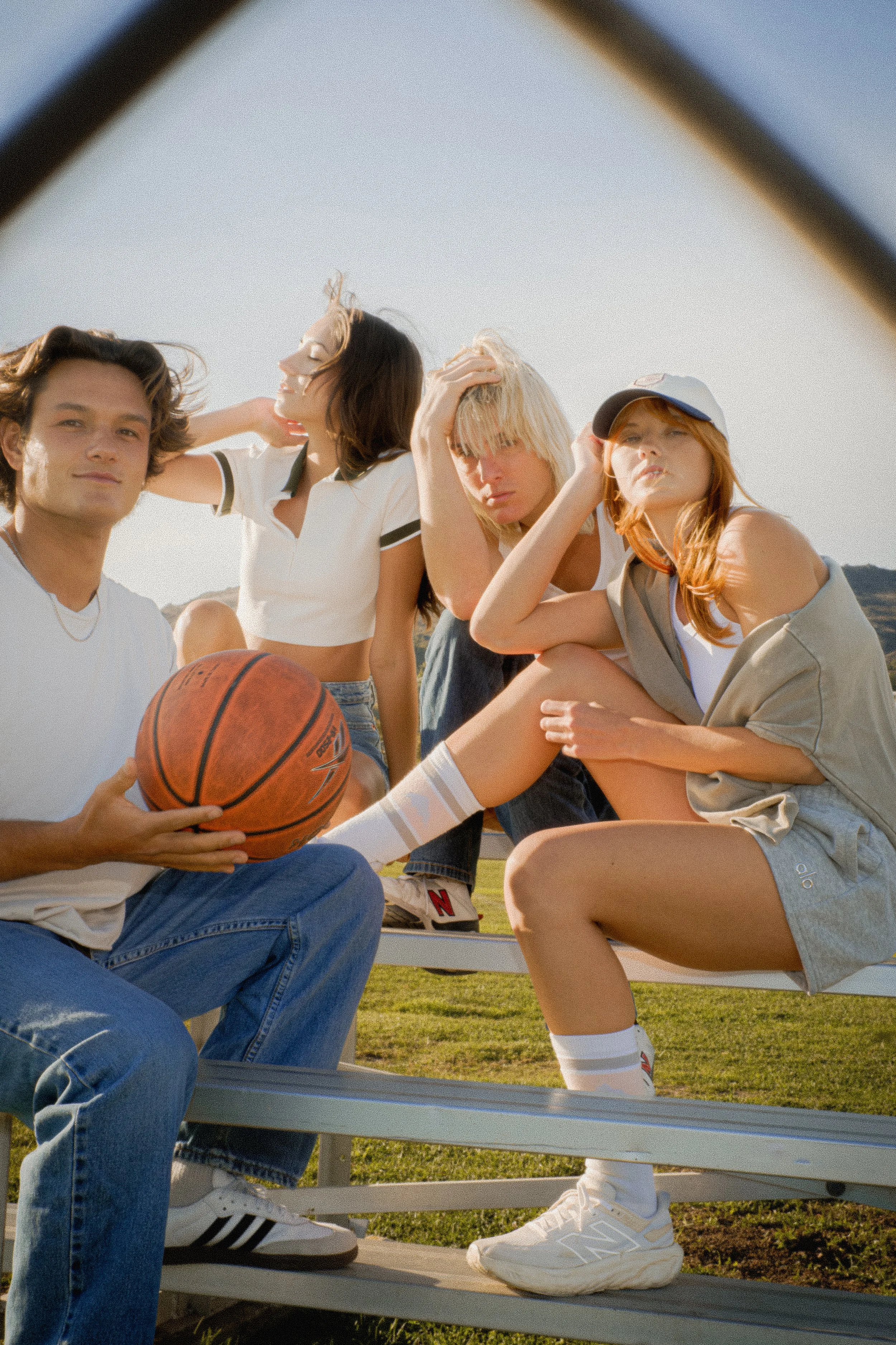 Group of four young people sitting on a park bench outdoors, with one holding a basketball, dressed in casual summer clothing, under a clear sky.