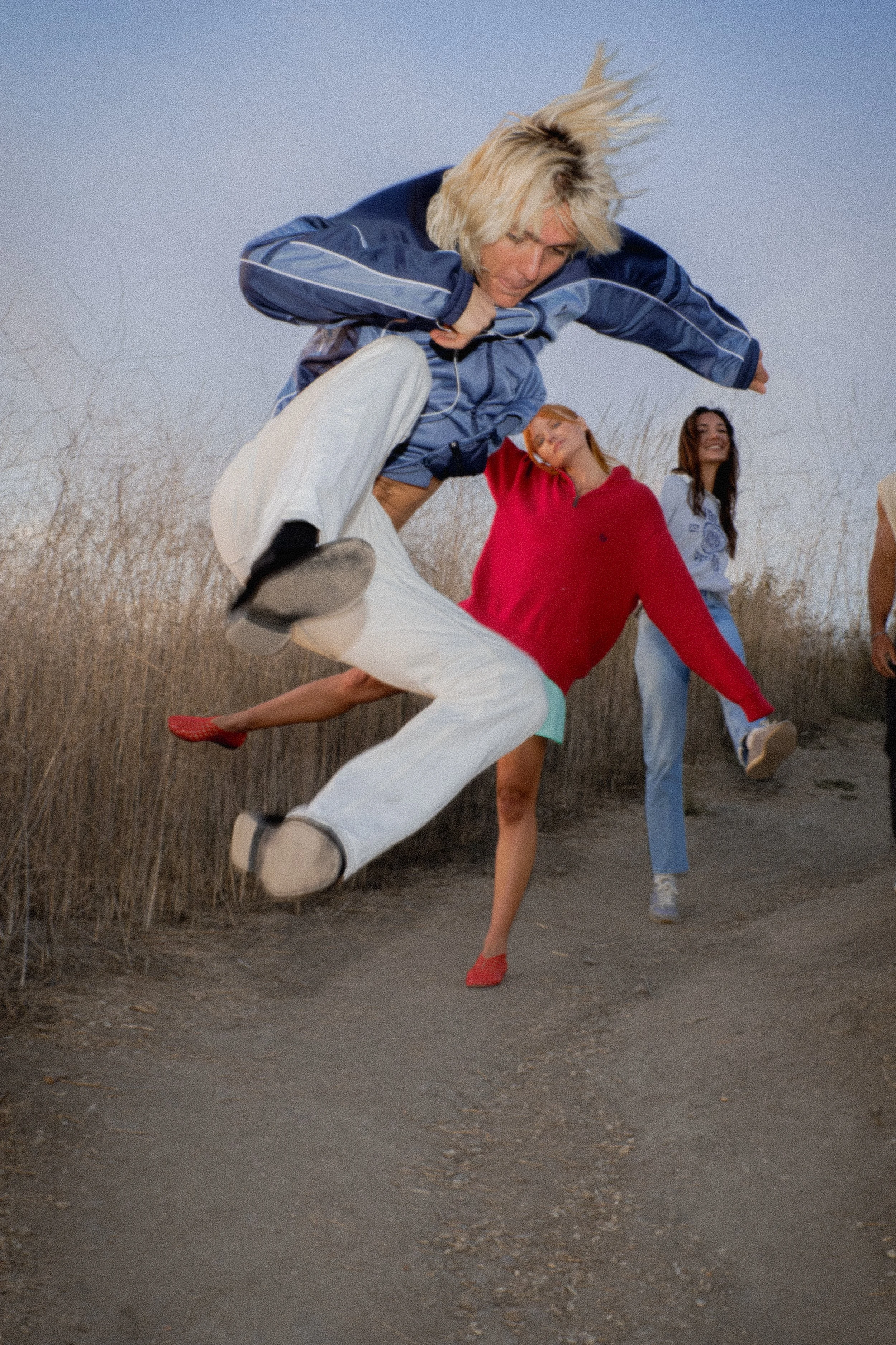 Young woman in a red sweater and white pants dancing in an outdoor setting with two other women in the background, one in a gray top and the other in a pink top. The woman in the foreground is balancing on one foot with her arms outstretched and one leg lifted behind her.