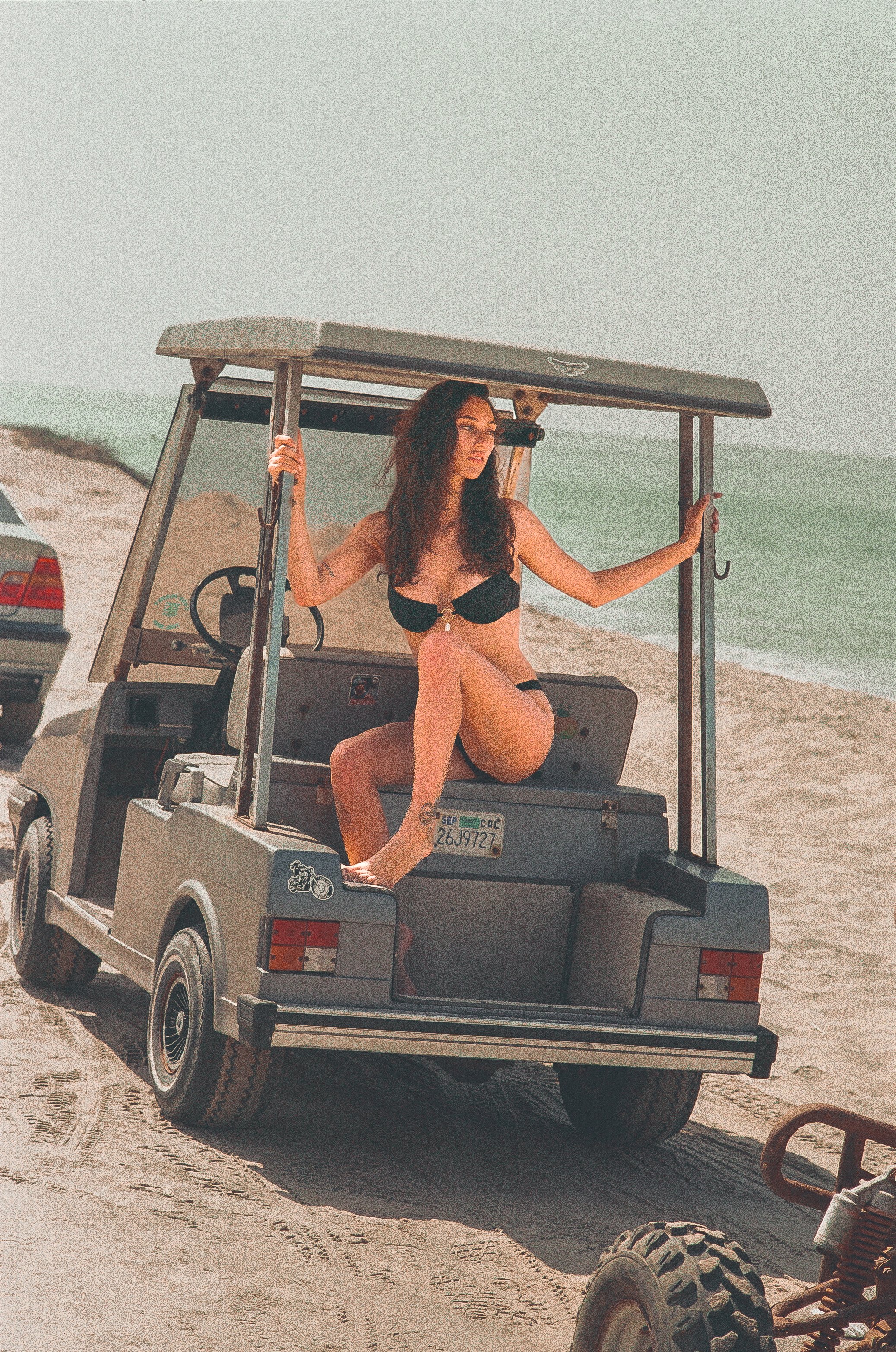 A woman in a black bikini with a bikini top and bottom, sitting on the back of a gray utility golf cart on a beach. She is holding onto the roof of the golf cart with one hand and looking off to the side. The beach has sand and the ocean is visible in the background.