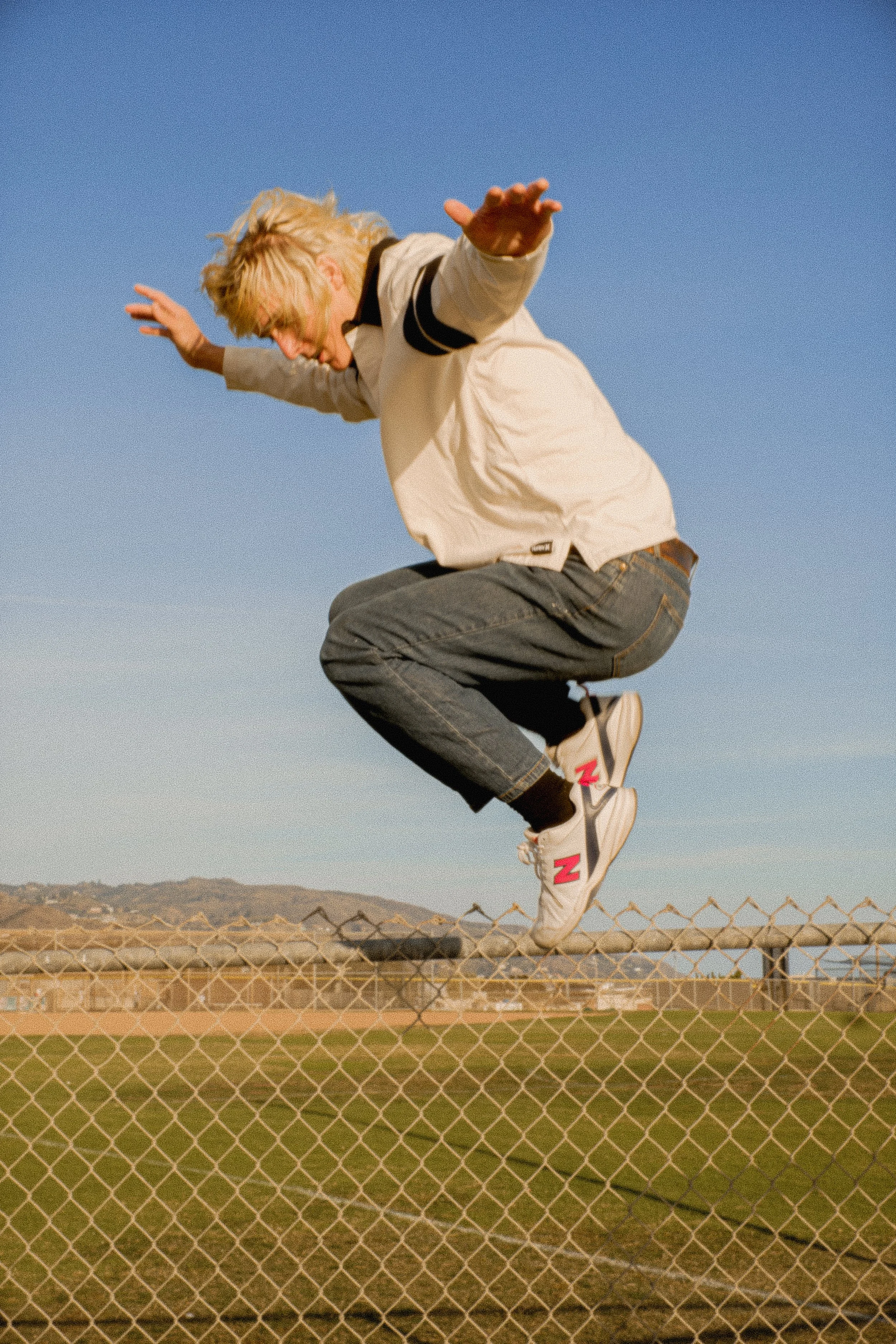 A person mid-jump over a chain-link fence at a sports field, wearing a cream jacket, jeans, and white sneakers with red "Z" logos, with a mountain range and clear blue sky in the background.