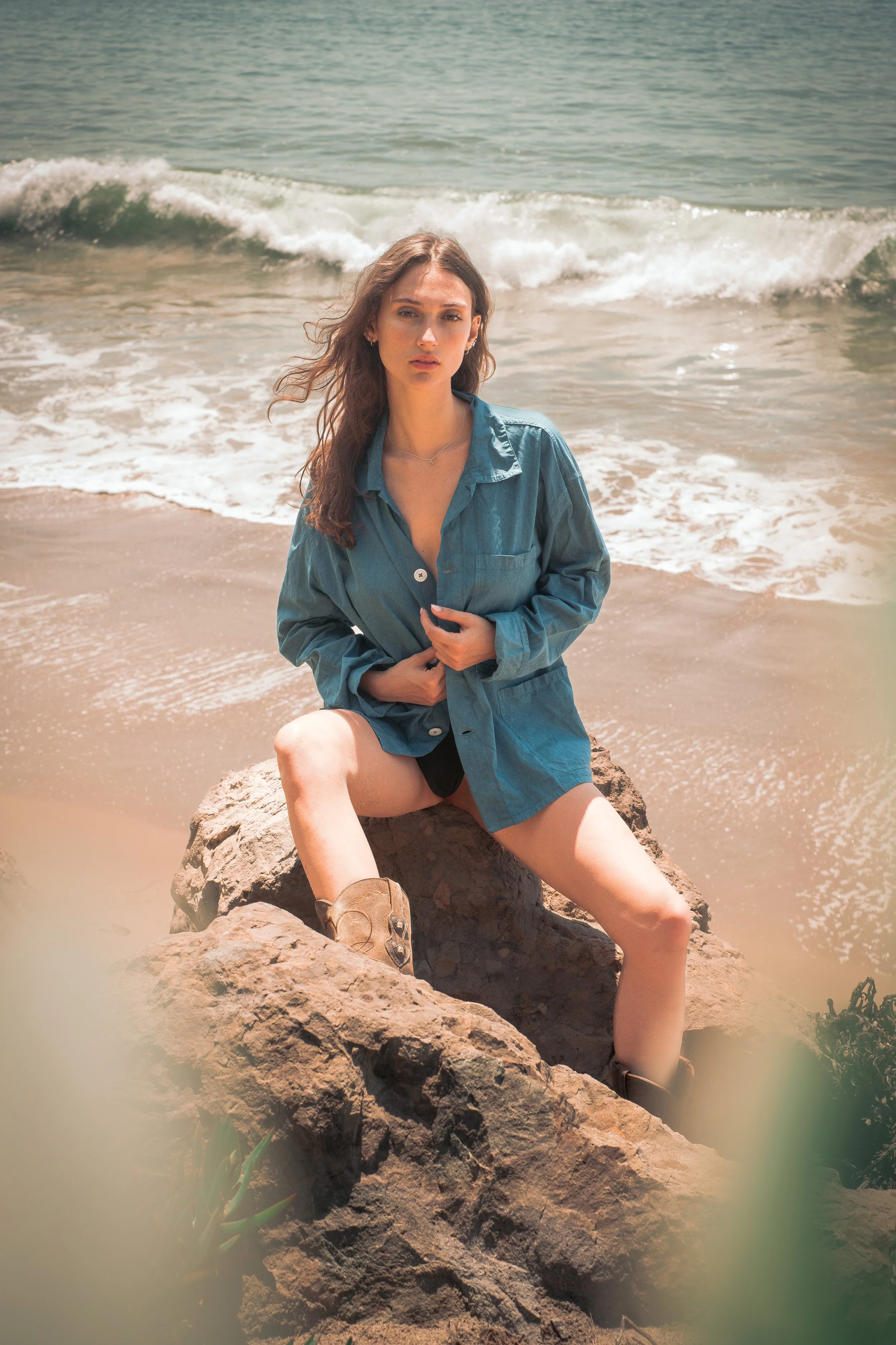 A young woman with wavy brown hair sitting on rocks at the beach, wearing a denim shirt and boots, with waves in the background.