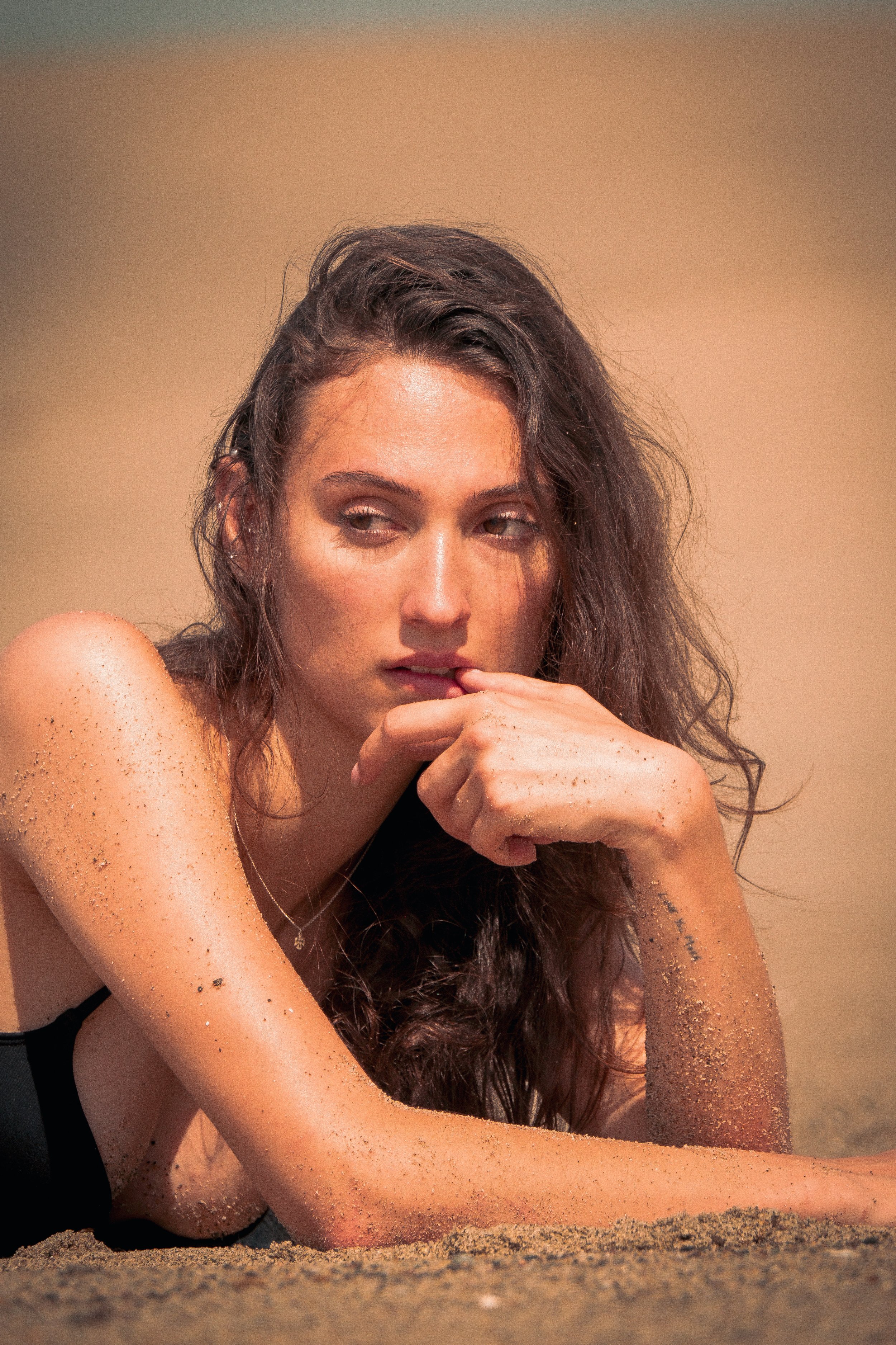Close-up of a woman with long brown hair, sitting on the beach with sand on her arms and shoulder, resting her chin on her hand.