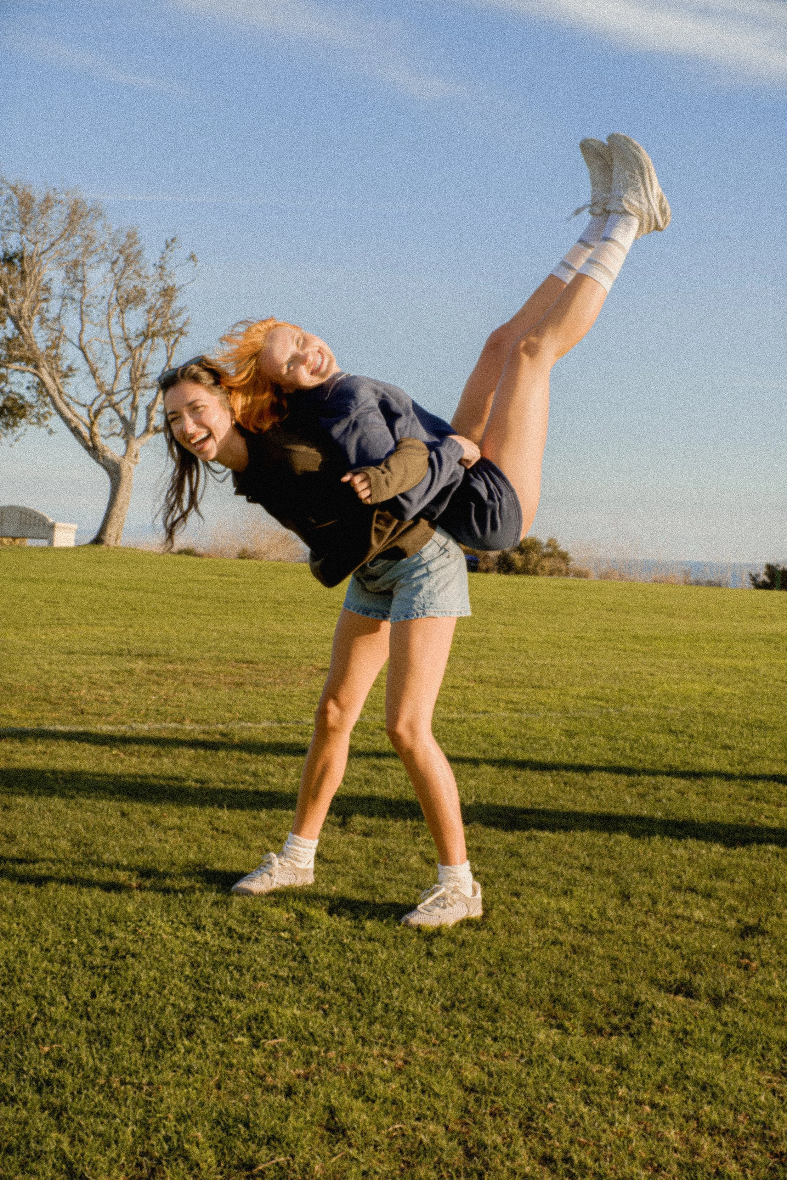 Two young women are having fun outdoors on a sunny day. One woman is lifting the other by her waist. Both women are smiling and laughing. They are on a grassy field with a tree and a bench in the background.