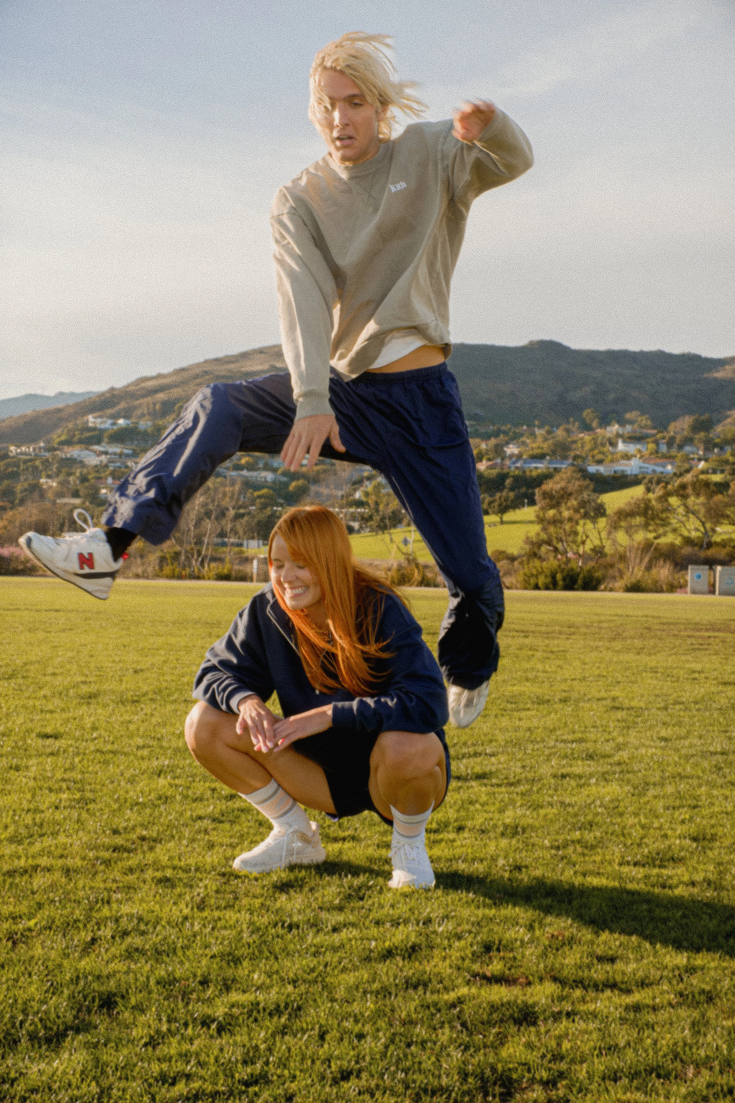 Two young women performing a human pyramid outdoors on a grassy field with hills and houses in the background during late afternoon or early evening.