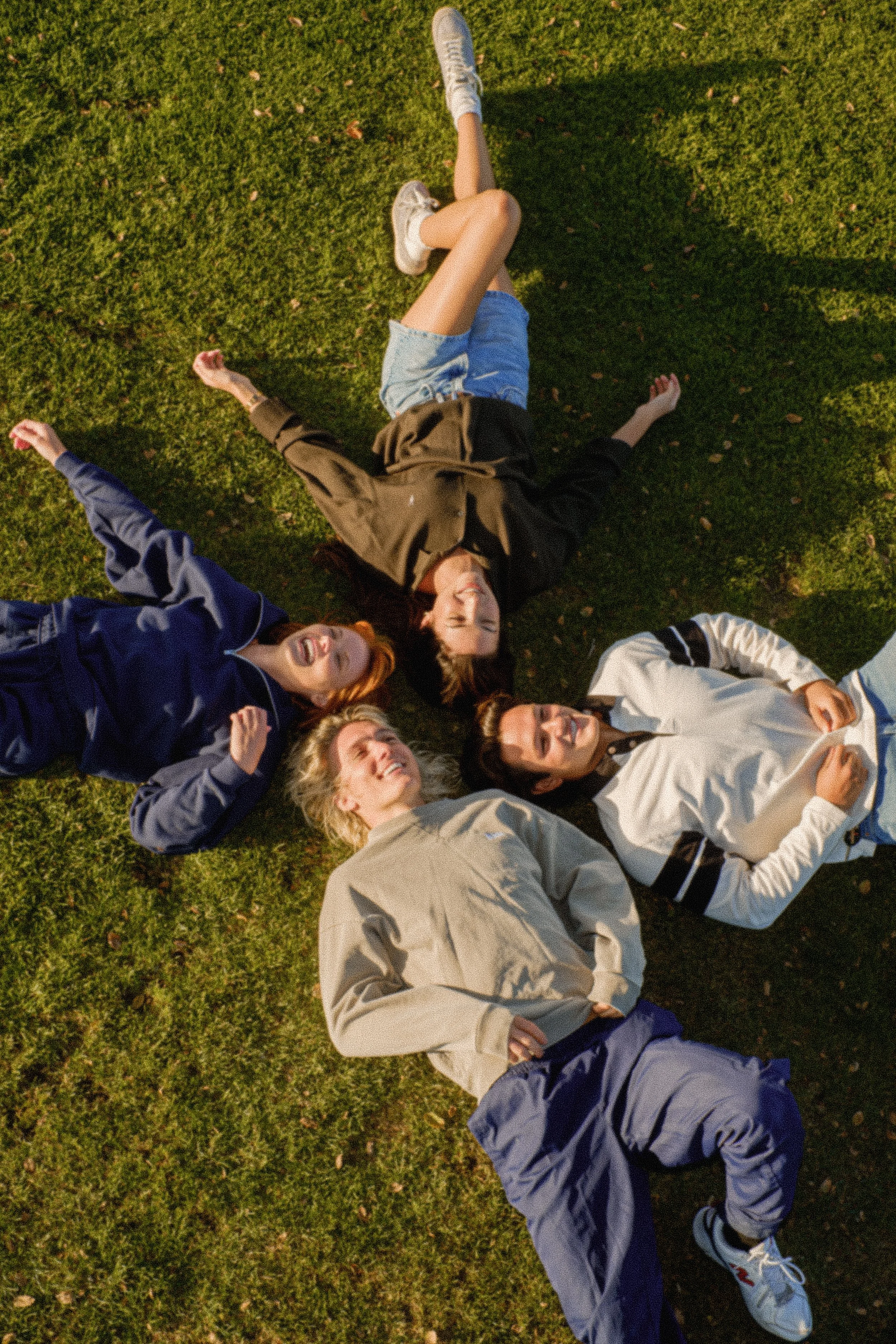 Group of young people lying on grass, looking up and smiling in outdoor setting.