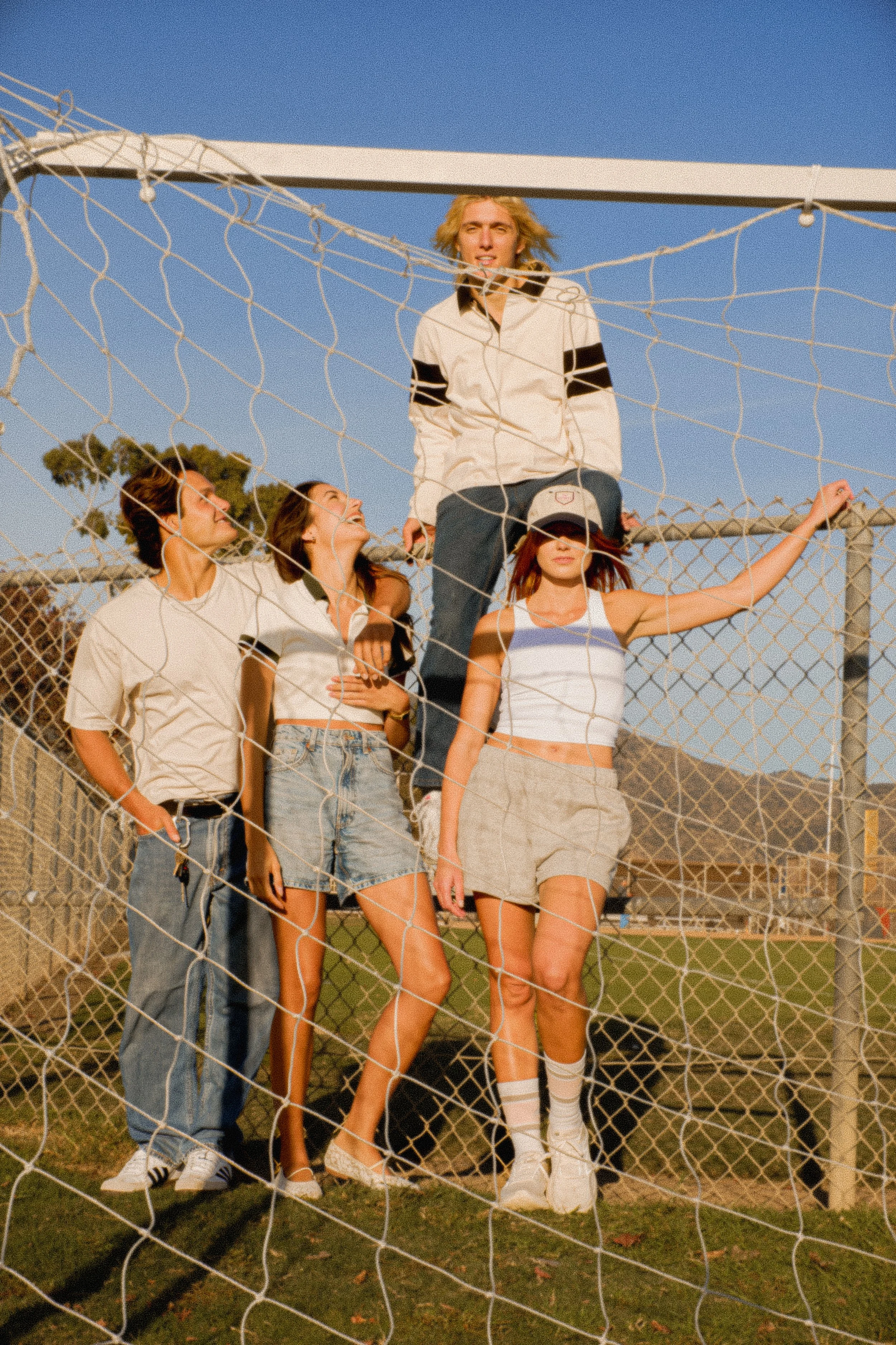 Group of five young adults standing behind a tennis or volleyball net on a grassy field with a clear blue sky in the background.
