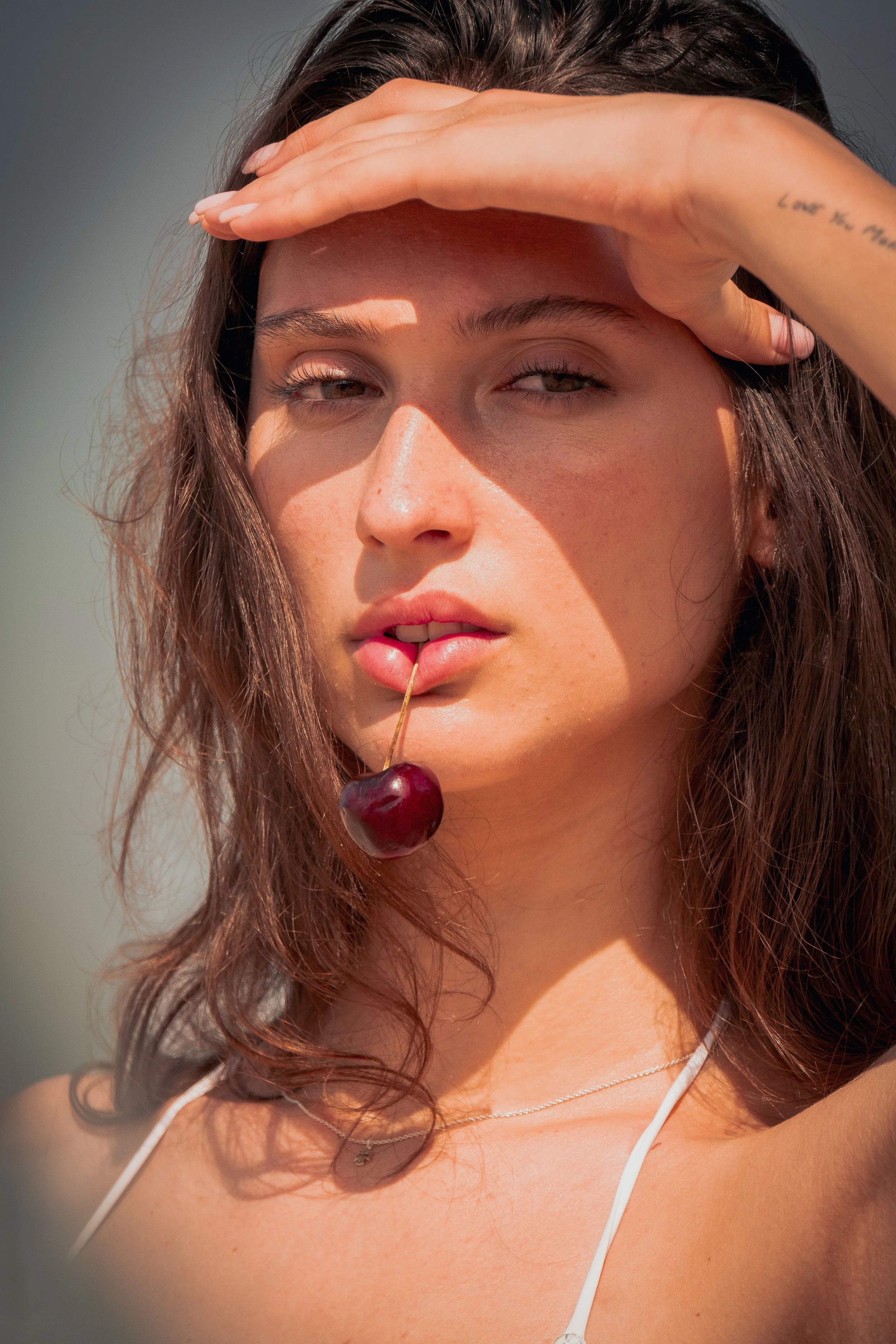 Close-up of a young woman with wavy brown hair shielding her eyes from the sun with her hand, holding a cherry in her mouth, wearing a thin necklace and white earbuds.