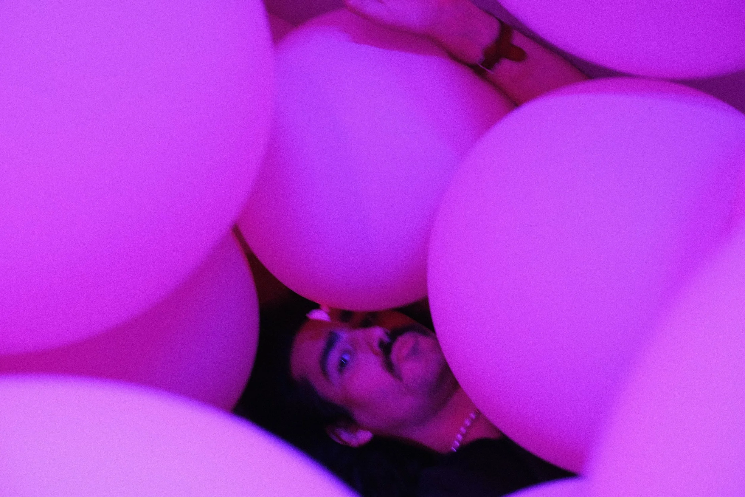 Close-up of pink balloons with a person smiling and making a peace sign in the background, illuminated by purple lighting.