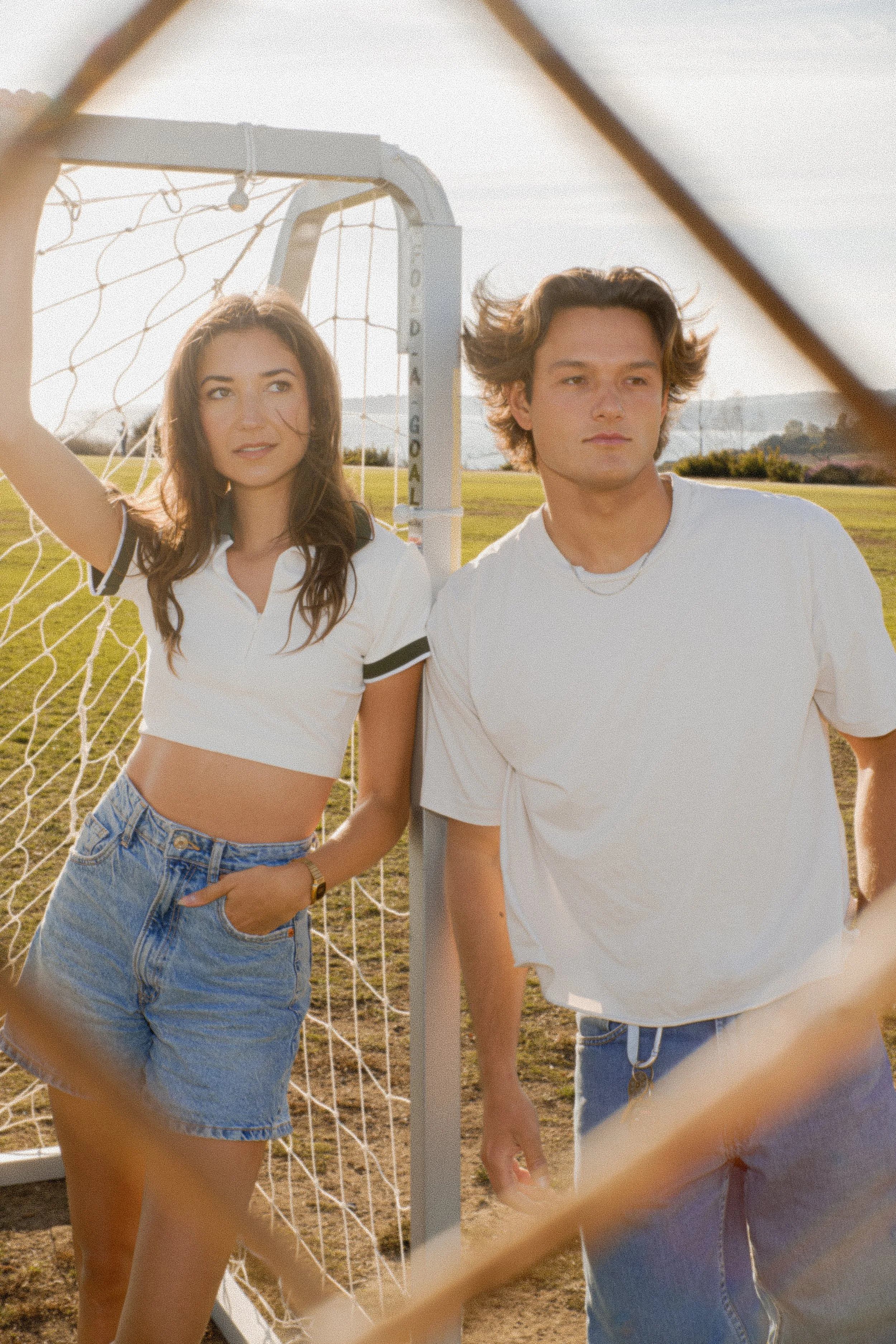 A young woman and a young man standing near a soccer goalpost outdoors on a sunny day, seen through a diamond-shaped chain-link fence.