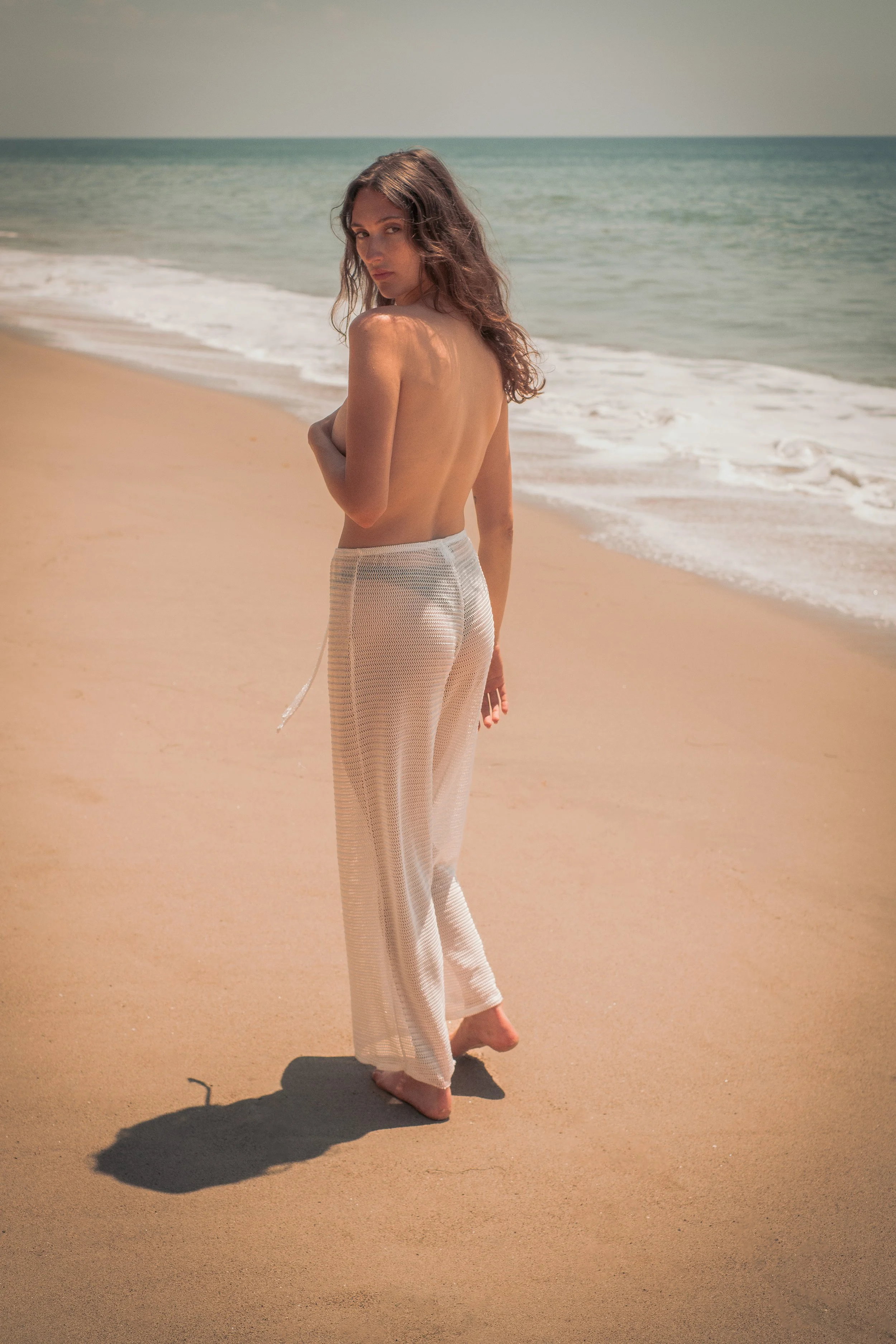 A woman with long, wavy brown hair stands barefoot on a sandy beach, facing the camera with her back partly turned. She appears to be topless, covering herself with her left arm. She wears loose, light-colored, wide-legged pants. The ocean with gentle waves is visible in the background under a clear, light sky.