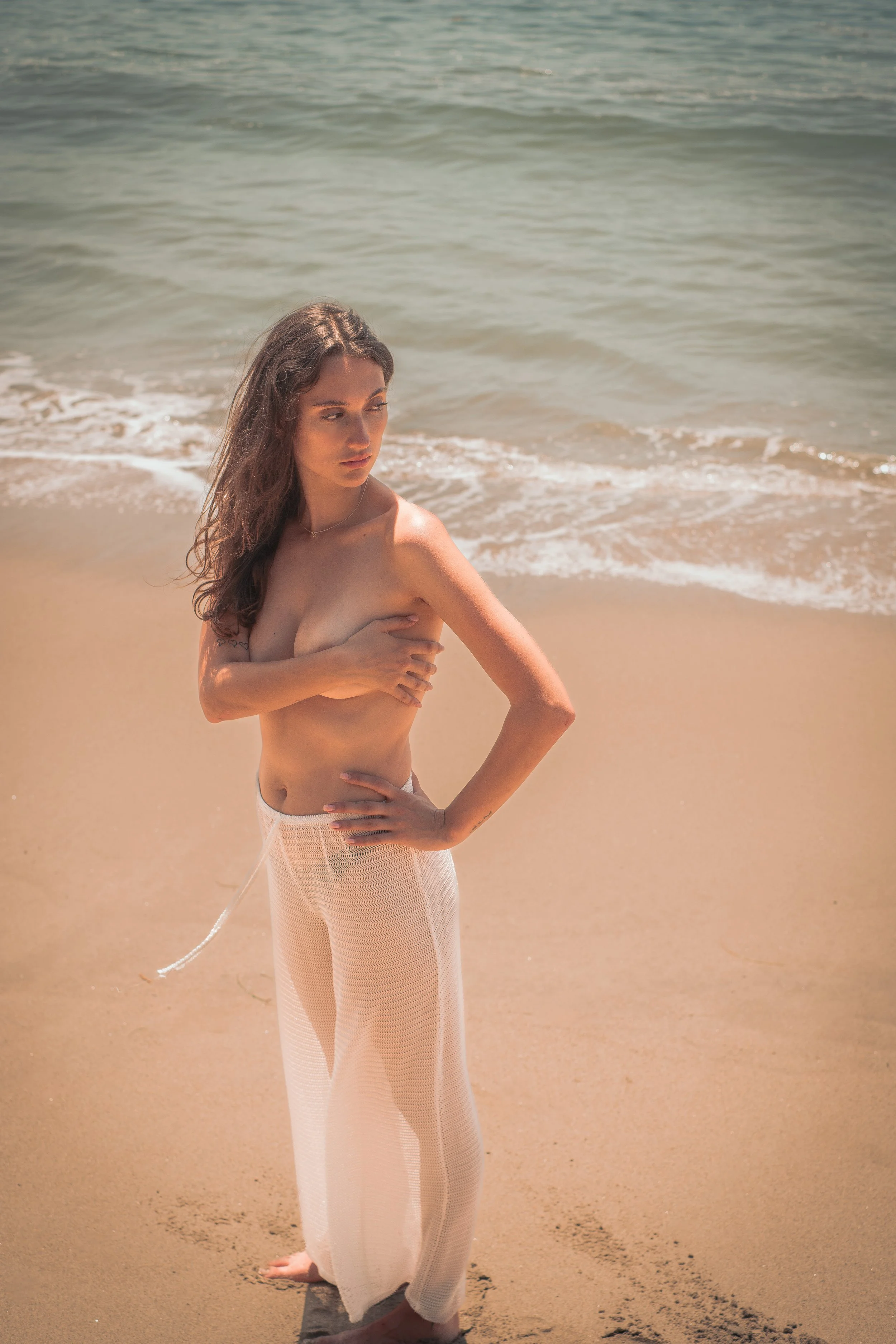 A woman standing barefoot on the sandy beach near the water, wearing light-colored, loose pants and covering her chest with one arm, with ocean waves in the background.