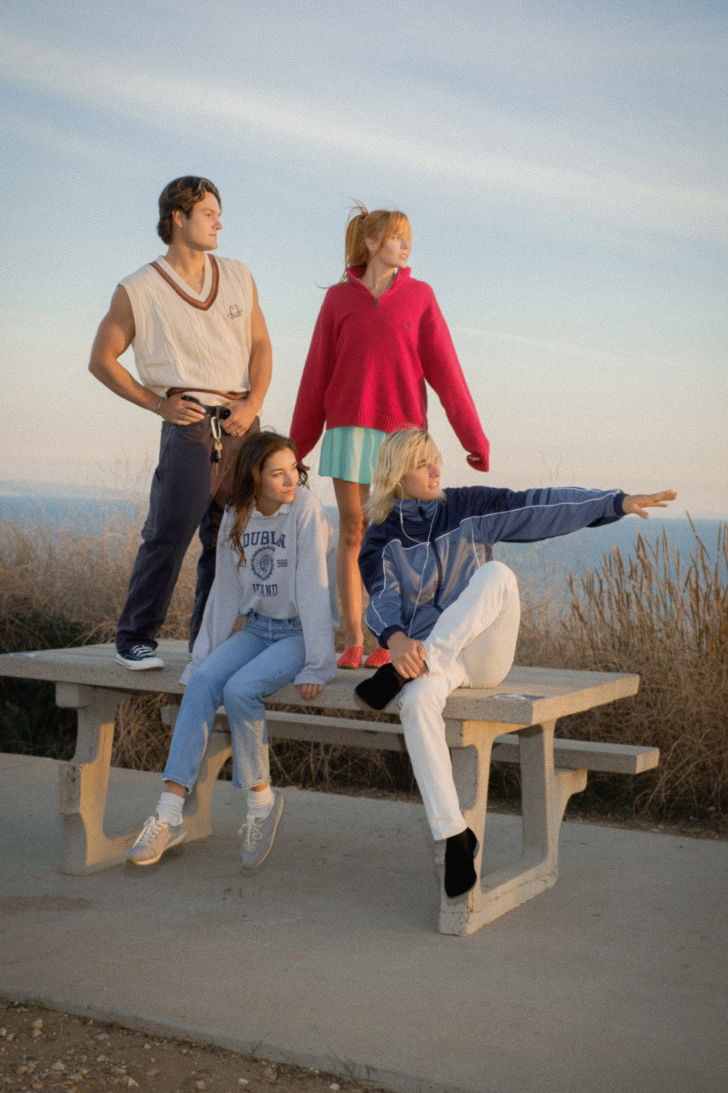 Group of five young people outdoors on a bench, with some standing and some sitting, looking off into the distance during sunset.