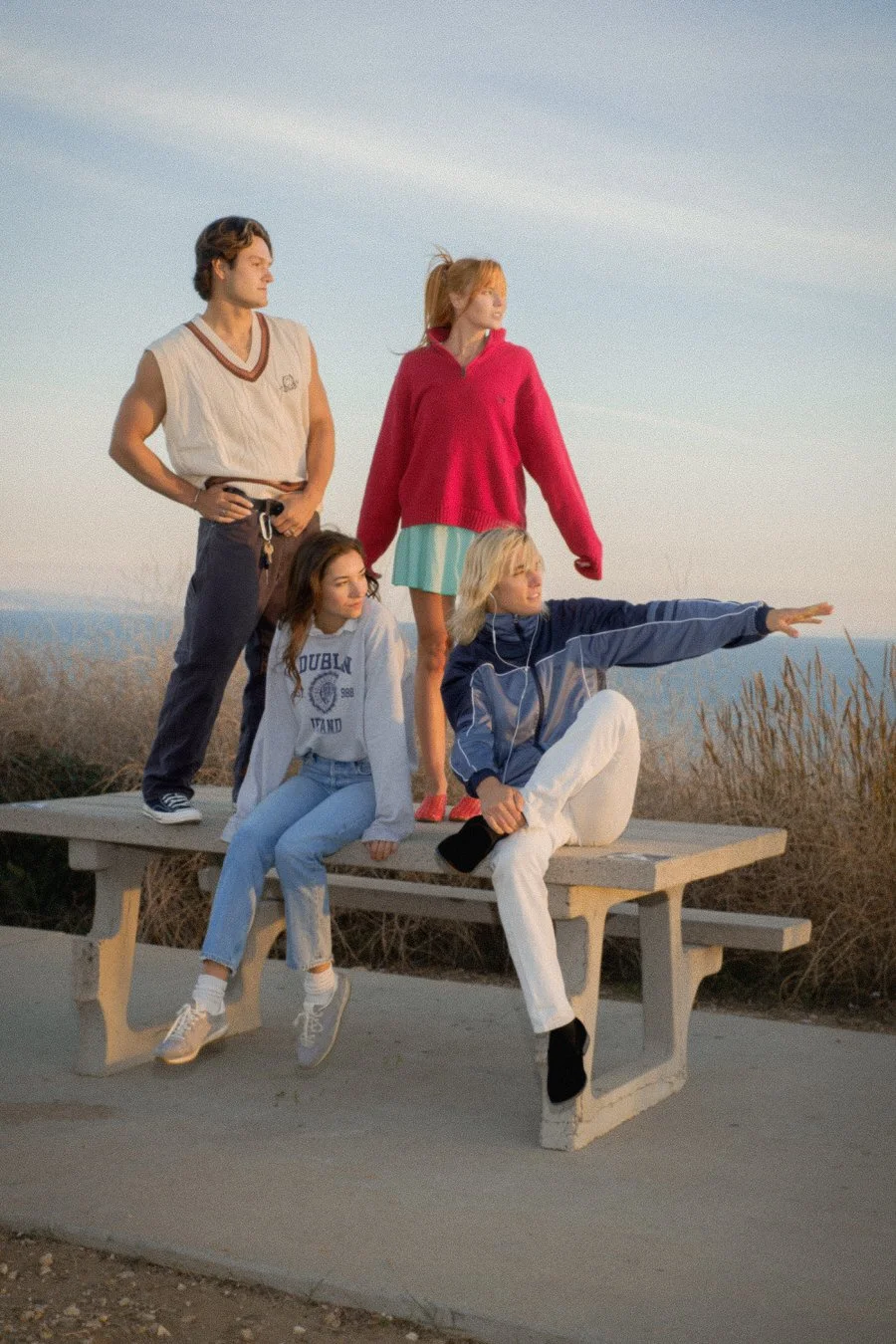 Four young women and one young man posing on a picnic table outdoors at sunset, with a scenic view of the ocean and sky in the background.