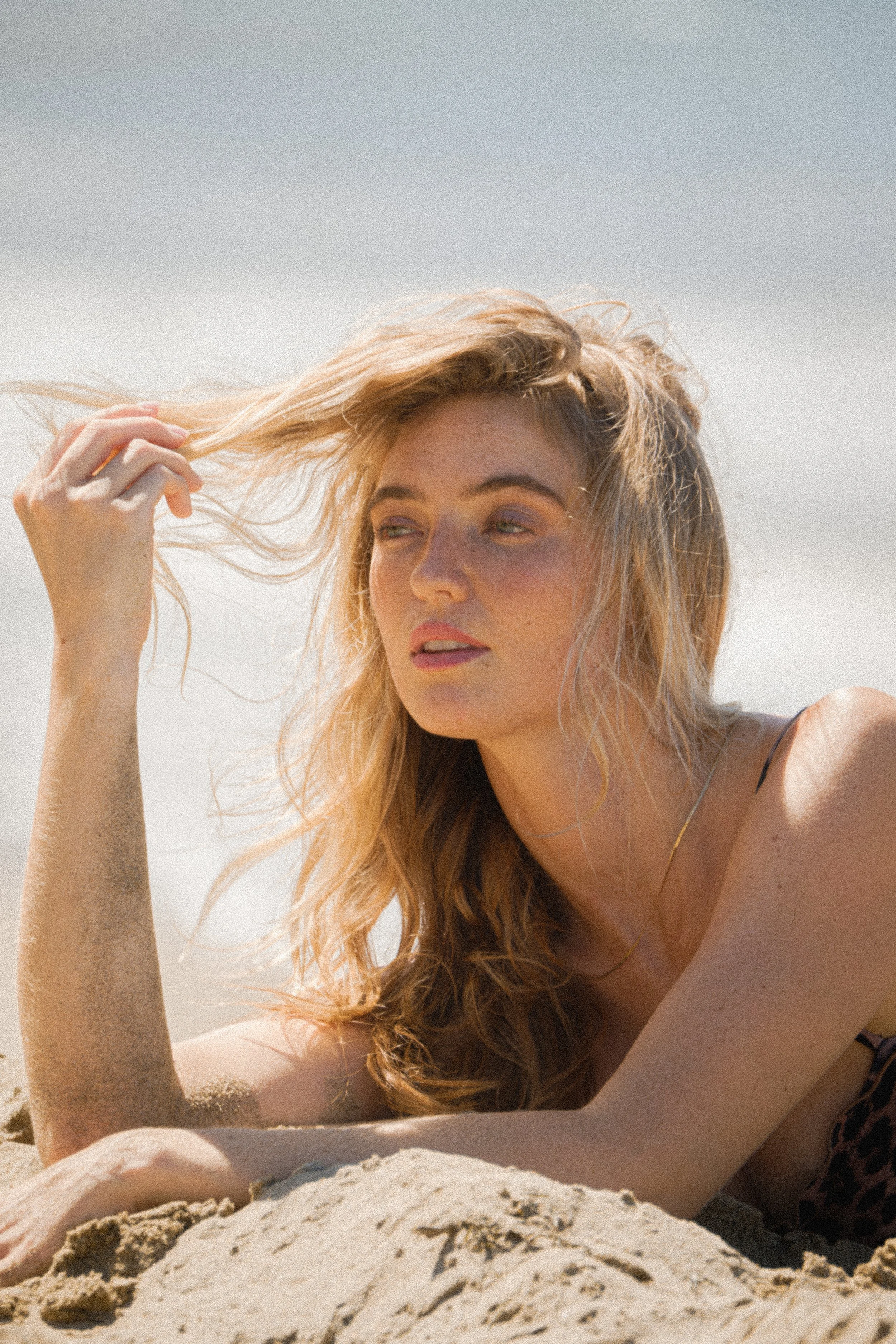 A woman with curly blonde hair lying on the sand at the beach, touching her hair with one hand, wearing a thin strap top and a delicate necklace.