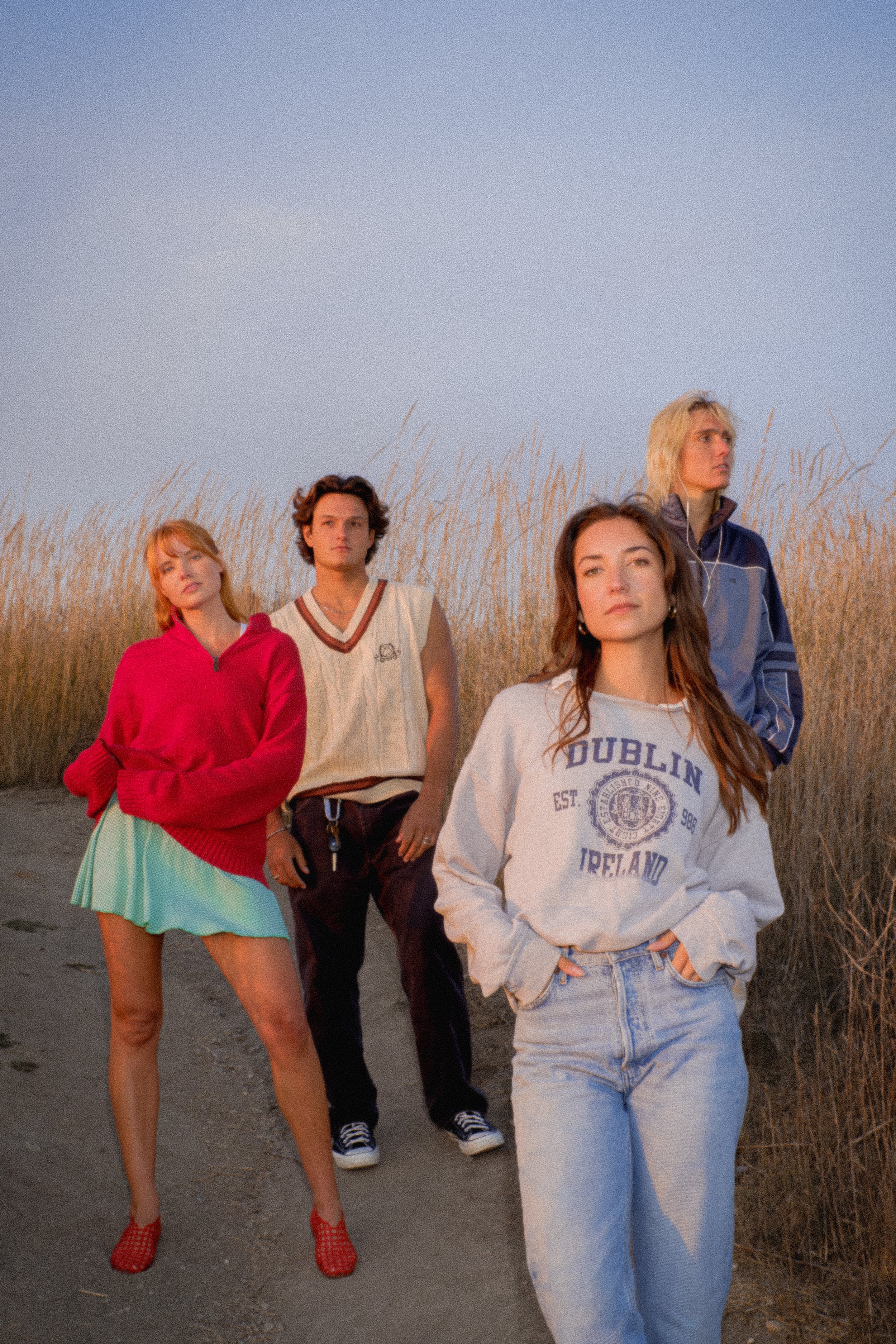 Four young adults standing outdoors on a dirt path with tall grass on either side, during sunset or sunrise. The group consists of two women and two men, wearing casual and sporty clothing, with serious or contemplative expressions.
