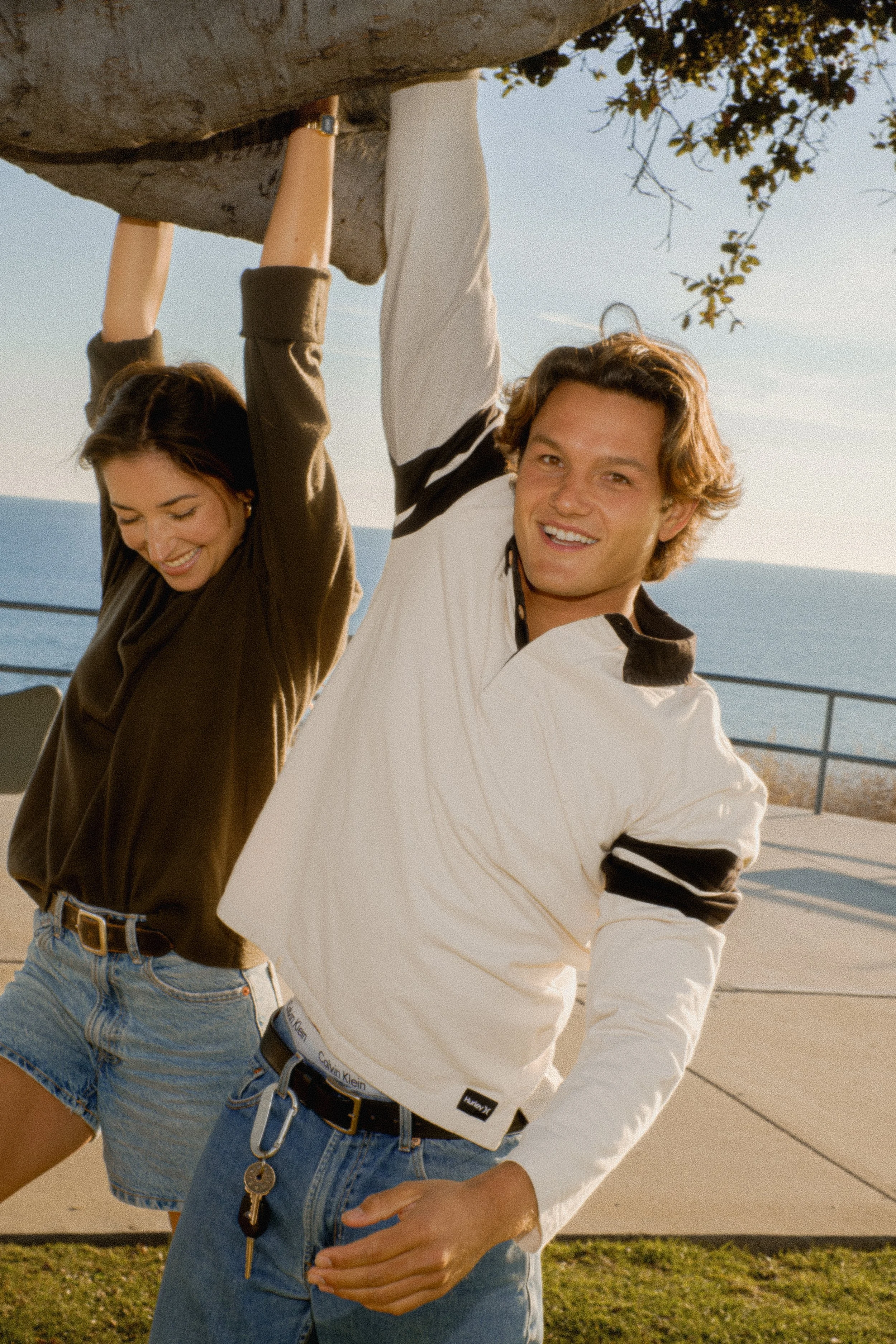 A young man and woman smiling on a sunny day; the man is holding onto a structure above them, and they are standing outdoors near a body of water.