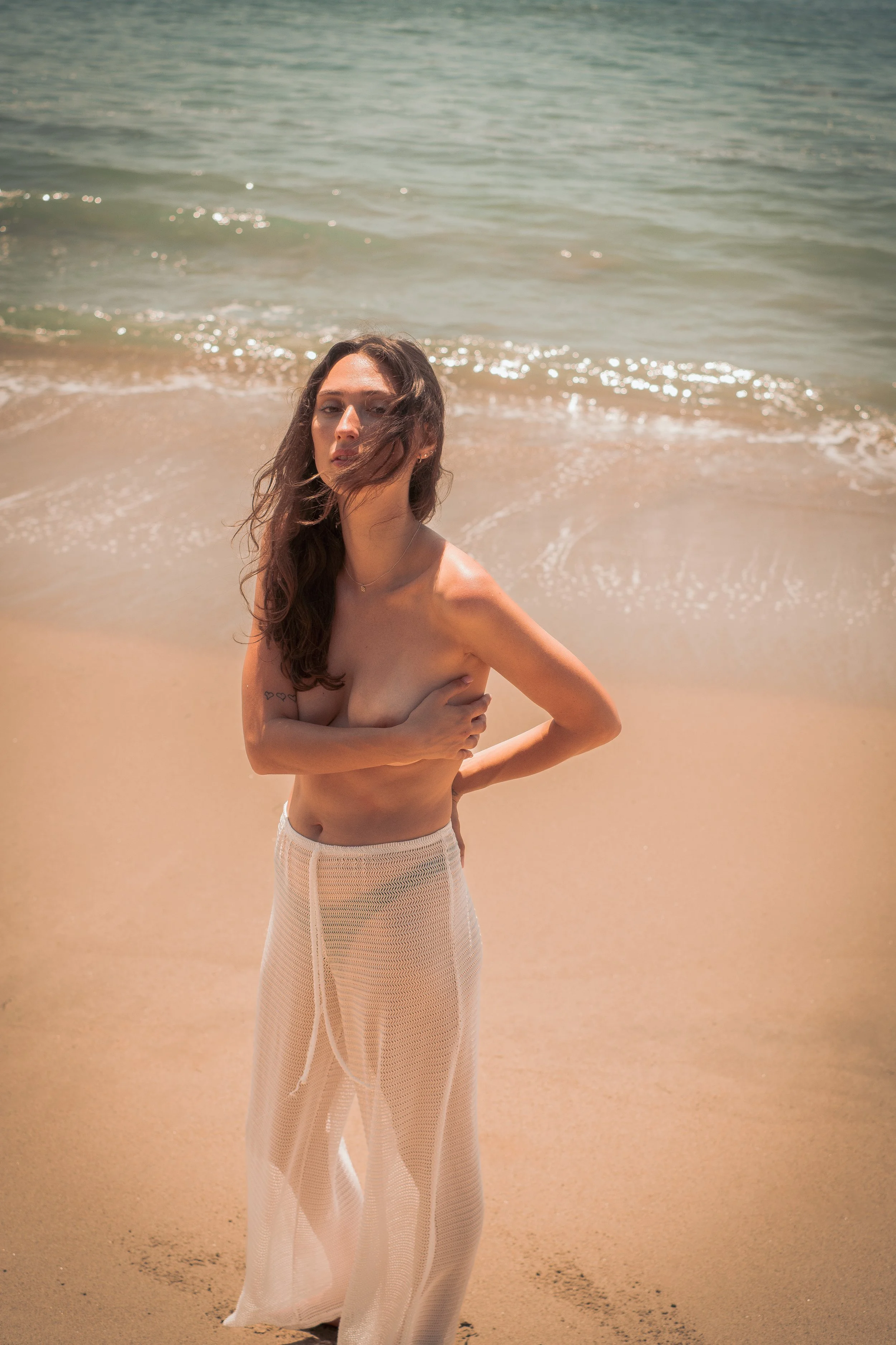 A woman standing on the beach near the water, wearing a sheer cream-colored skirt, with long brown hair, and holding her arm across her chest.